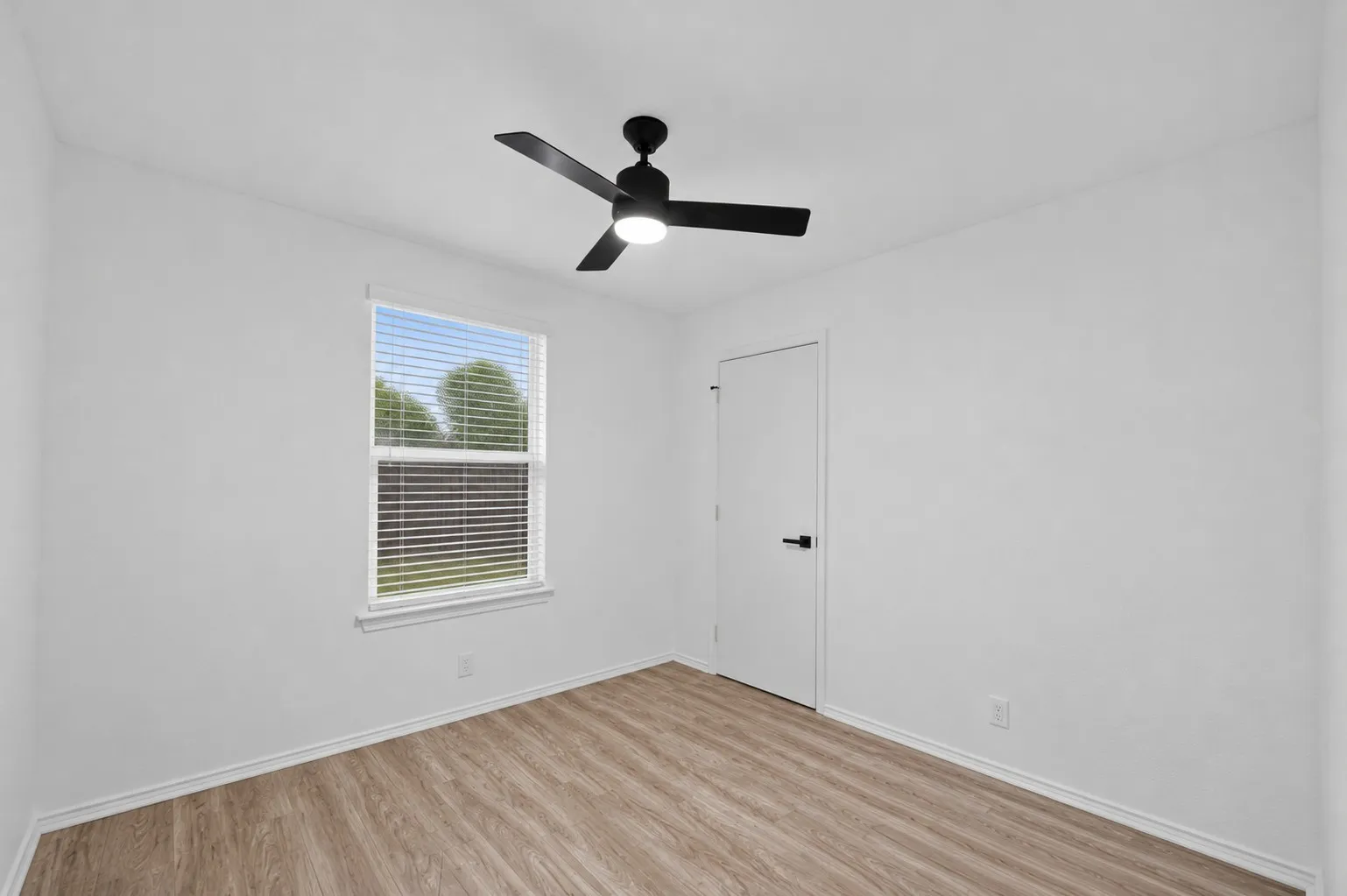 Bright room featuring a modern black ceiling fan with integrated lighting, wood-finish flooring, a white trim window with blinds, and white walls