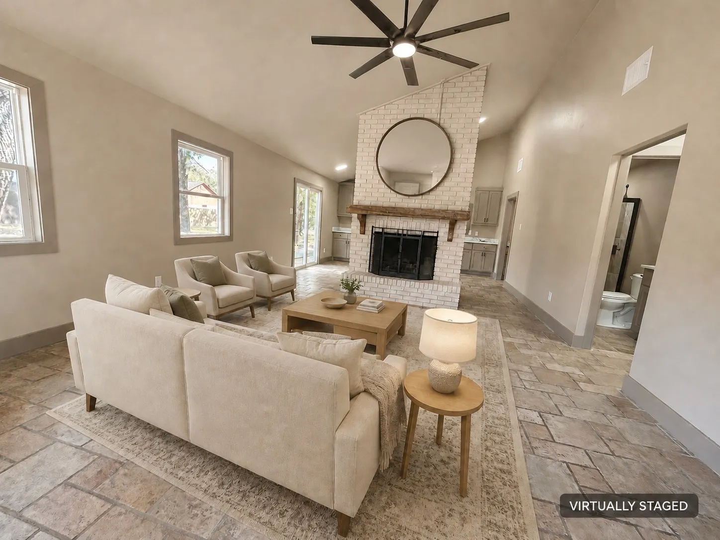 Vaulted ceiling, stone-look flooring, white brick fireplace with wood mantel, ceiling fan, and sliding glass doors