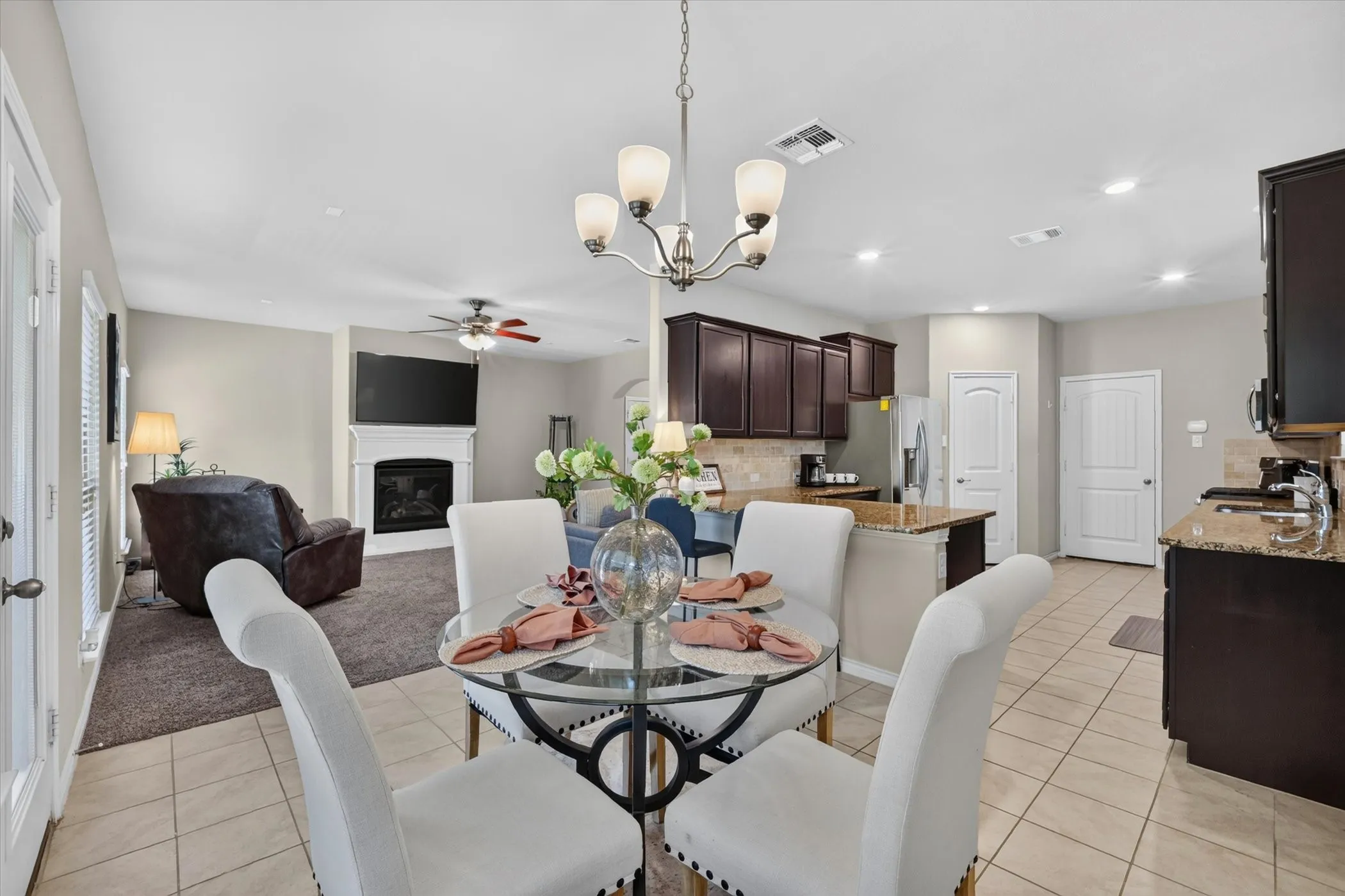 Open-concept layout featuring a dining area with a contemporary chandelier, a kitchen with dark wood-finish cabinetry and granite countertops, and a living area with a fireplace and ceiling fan