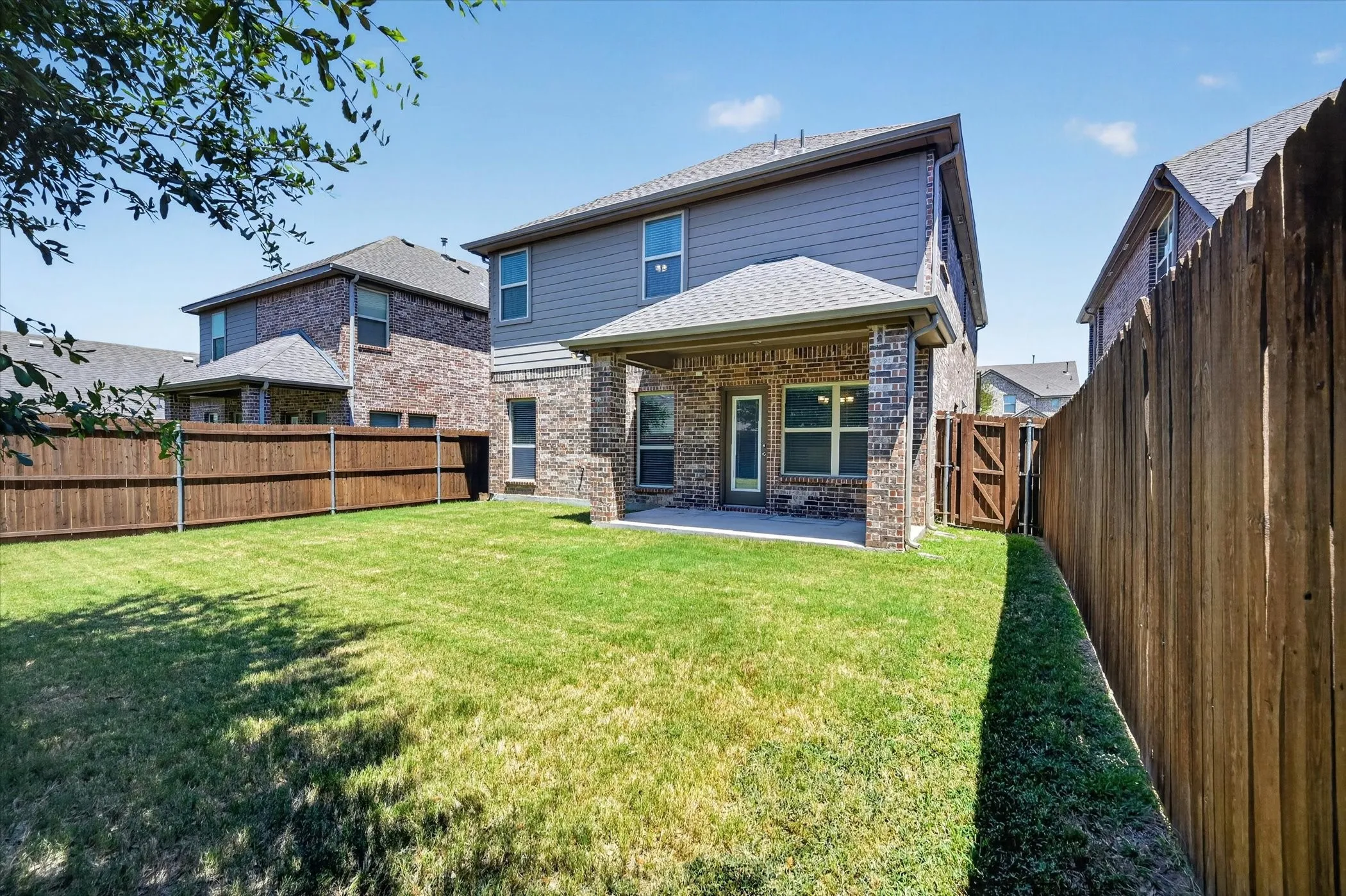 Expansive rear lawn featuring a covered patio with brick and stone columns, full privacy fencing, and a side access gate