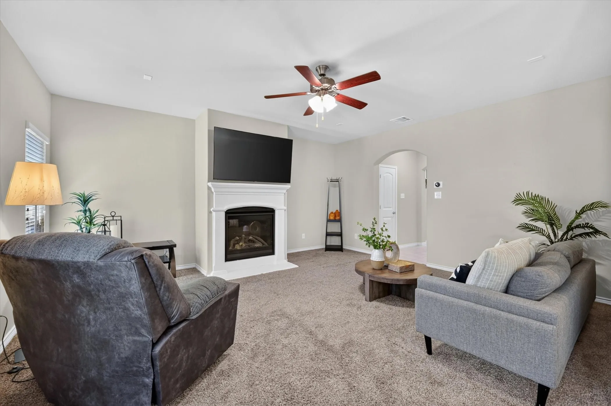 Living room featuring a gas fireplace with a white mantel, light gray walls, brown carpet flooring, and a ceiling fan with light fixture