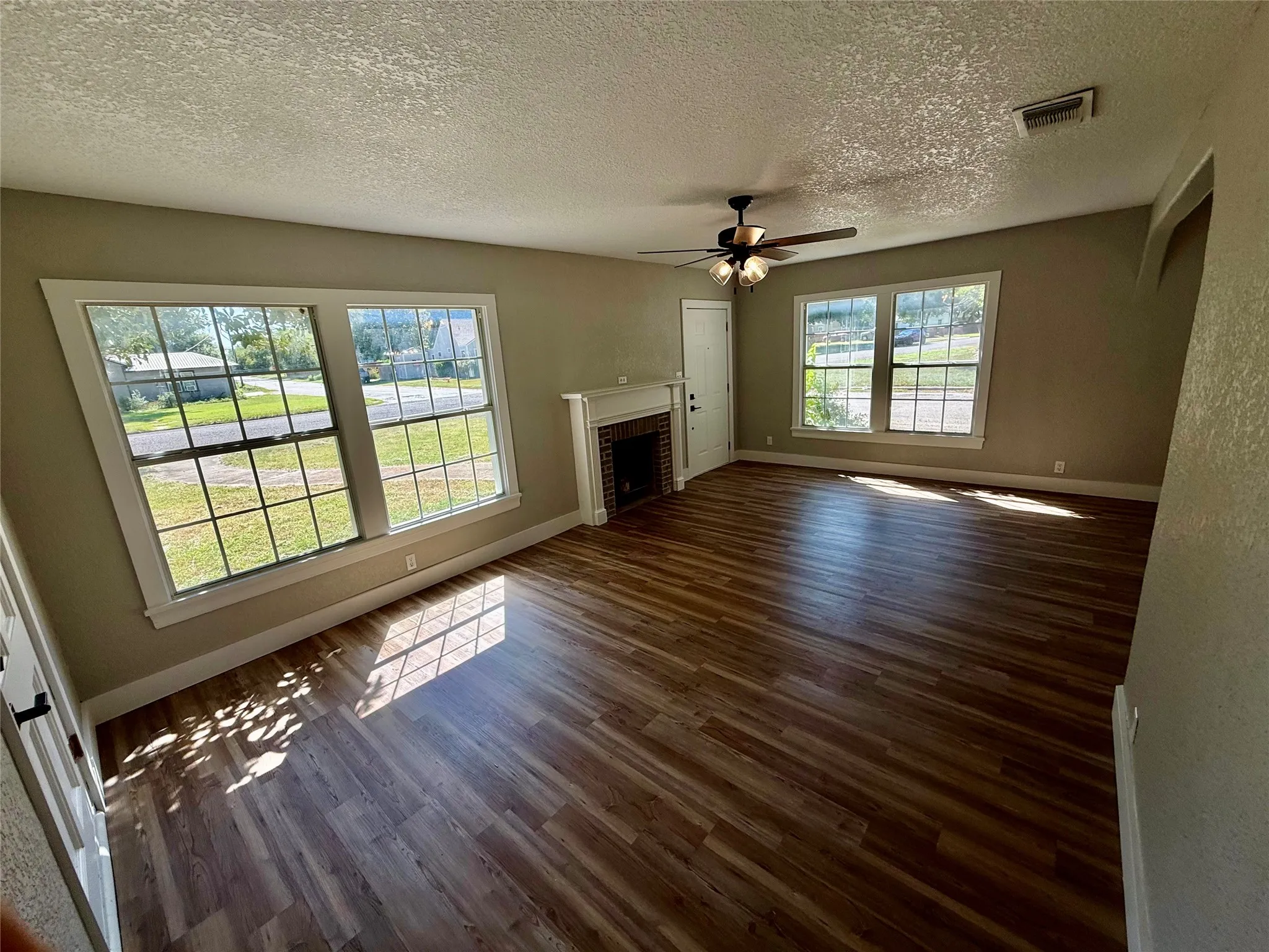 Spacious living area featuring wood-finish flooring, multiple windows, a ceiling fan, and a fireplace with a white mantel