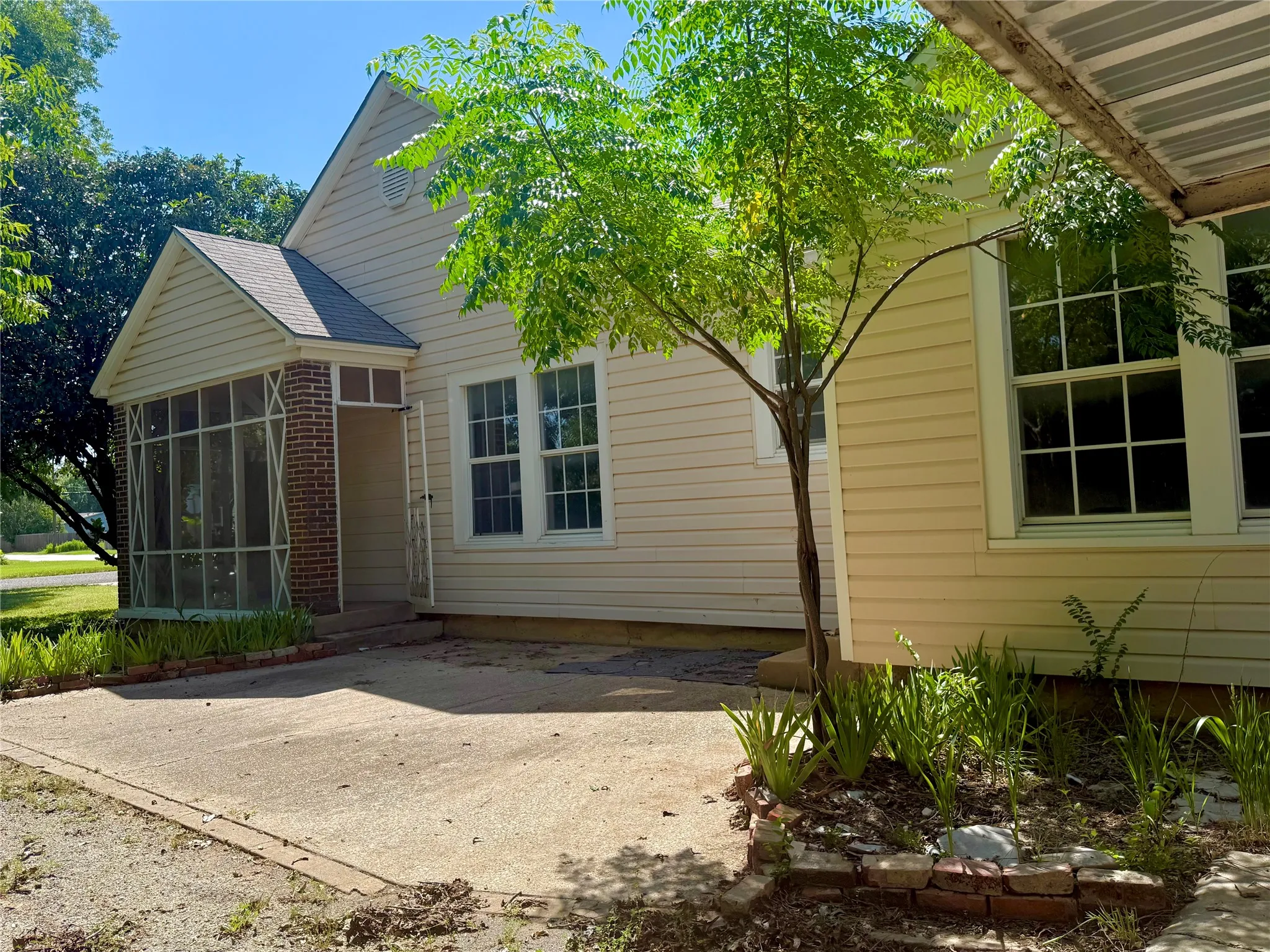 Cream-colored siding exterior featuring a covered porch with brick pillars and screening, multiple double-hung windows, and a mature tree