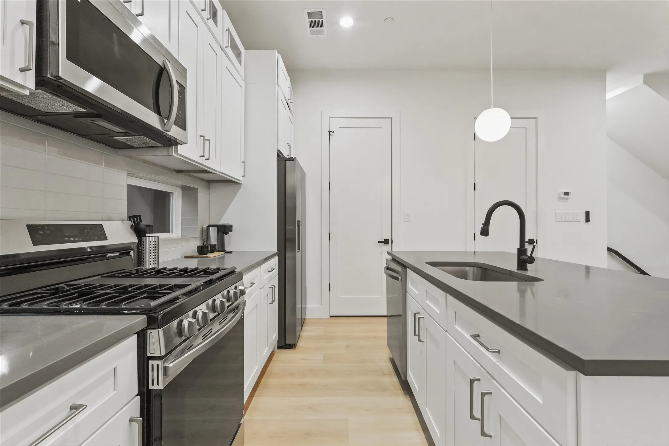 Kitchen with stainless steel appliances, light wood finished floors, white cabinets, hanging light fixtures, and an island with sink