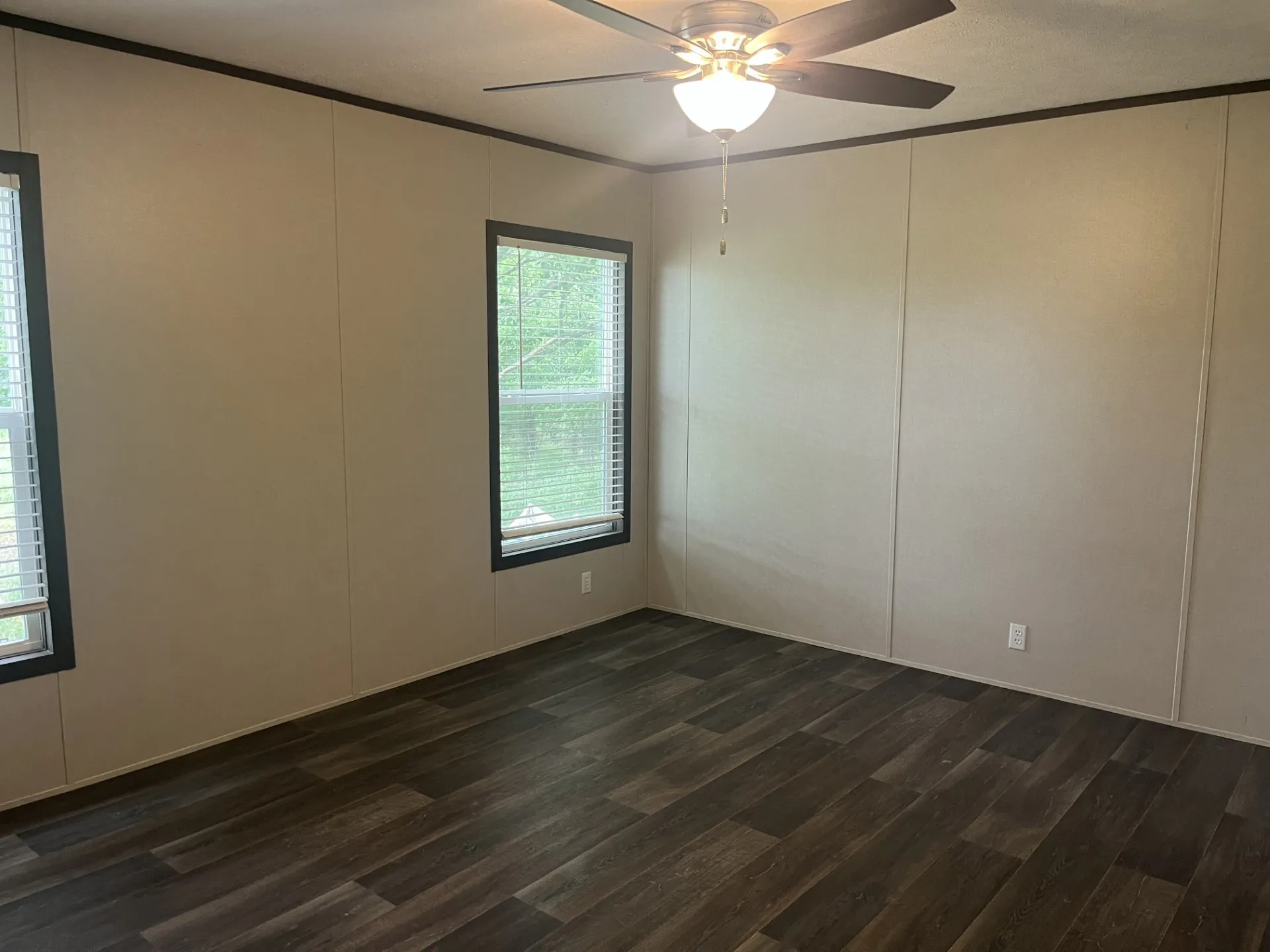 Room featuring wood-finish flooring, two windows with blinds, a ceiling fan with integrated lighting, and neutral wall panels with dark trim