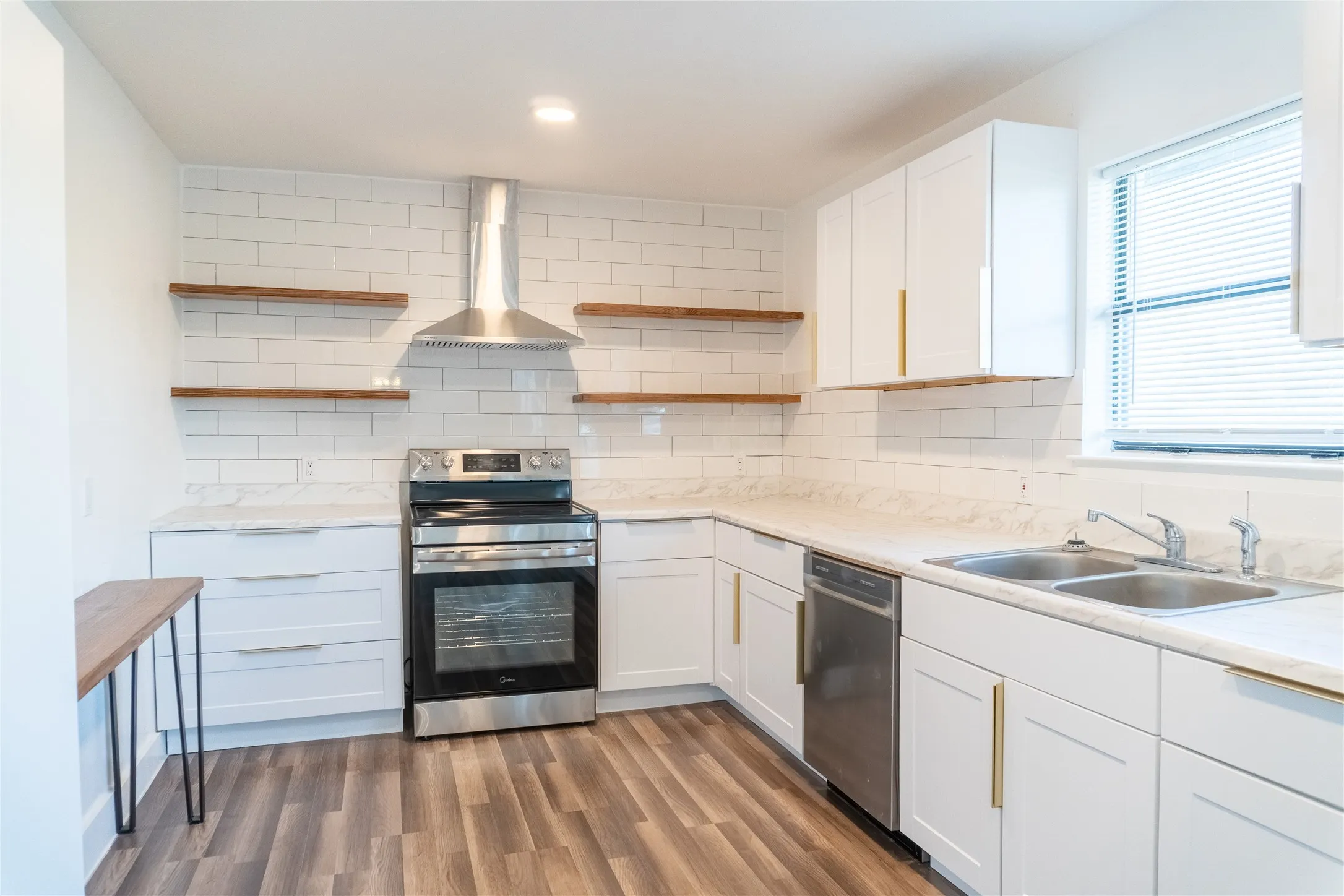 Updated kitchen featuring wood-finish flooring, white cabinetry with gold-tone hardware, stainless steel appliances, a subway tile backsplash, and light-toned countertops