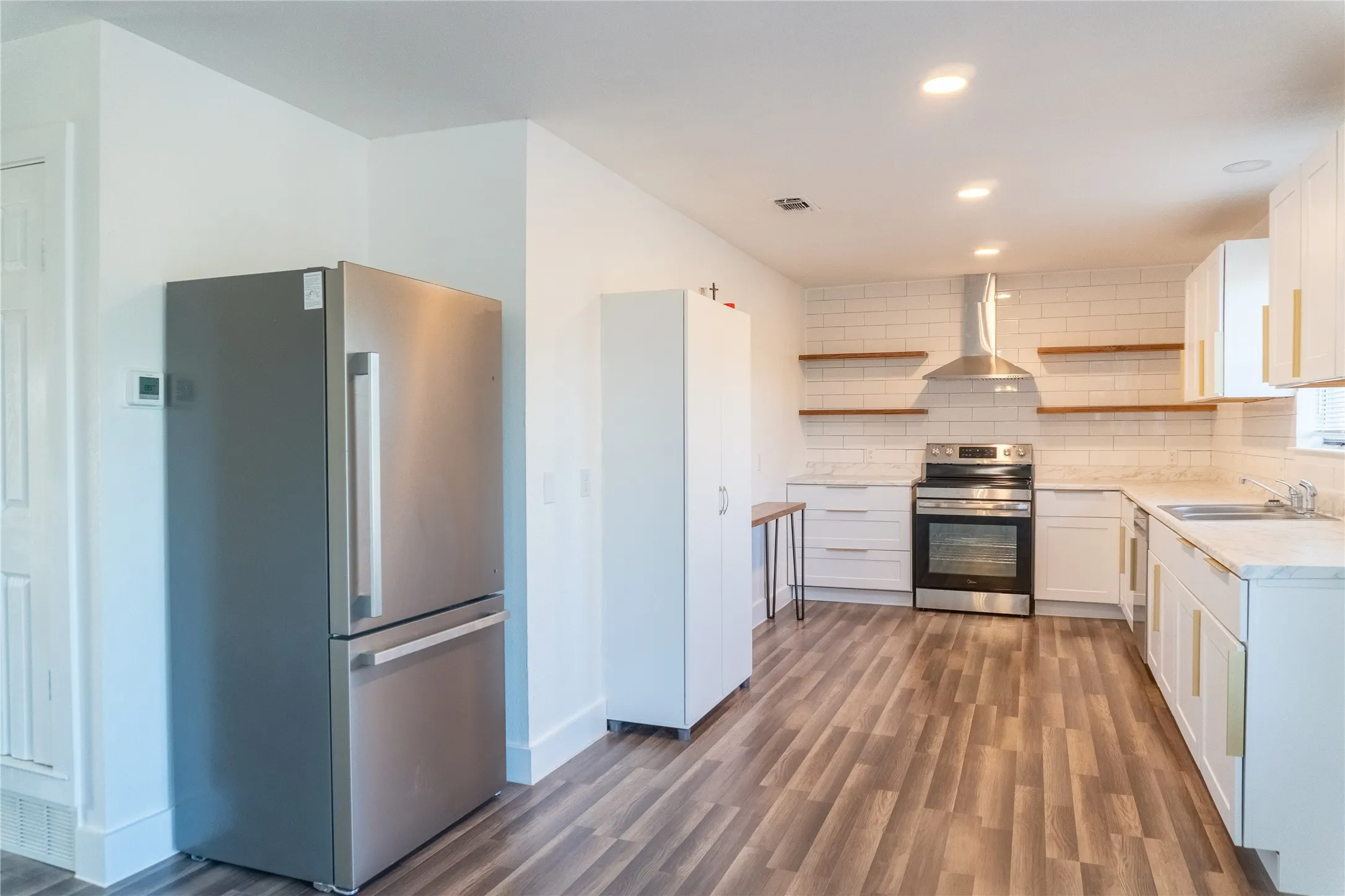Open-concept kitchen featuring wood-finish flooring, white cabinetry with gold-tone hardware, a stainless steel appliance suite, white subway tile backsplash, and recessed lighting