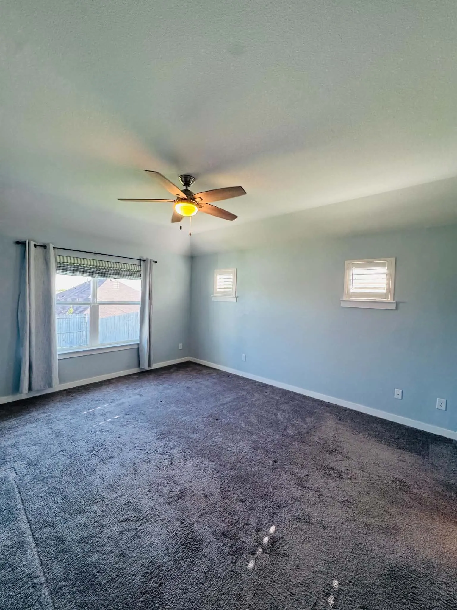 Expansive carpeted room featuring a ceiling fan with integrated lighting, multiple windows with blinds and curtains, and light blue wall finishes