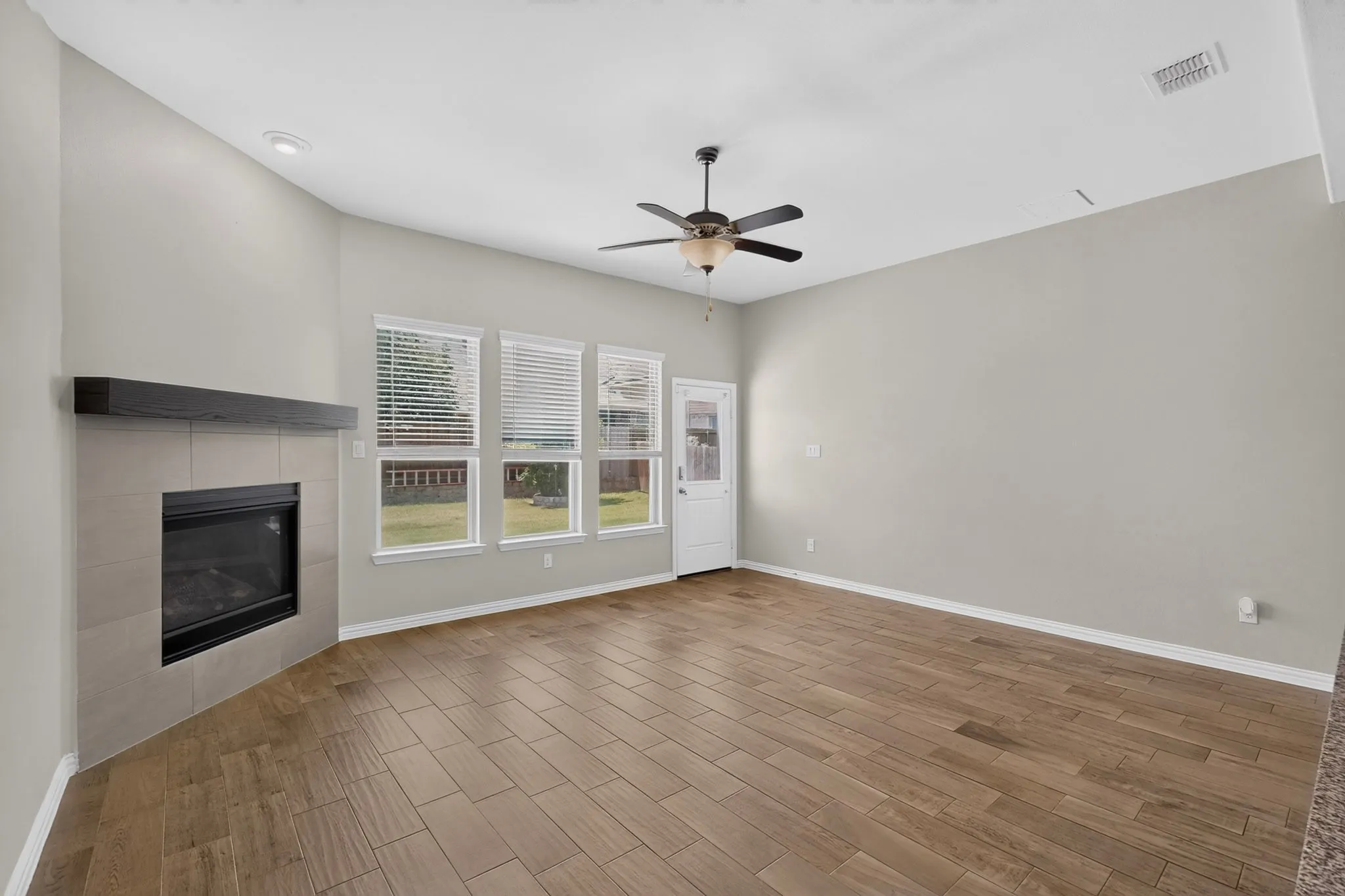 Spacious room featuring wood-finish tile flooring, a corner fireplace with a dark wood mantel and light-toned tile surround, multiple windows with blinds, a ceiling fan, and a white exterior door