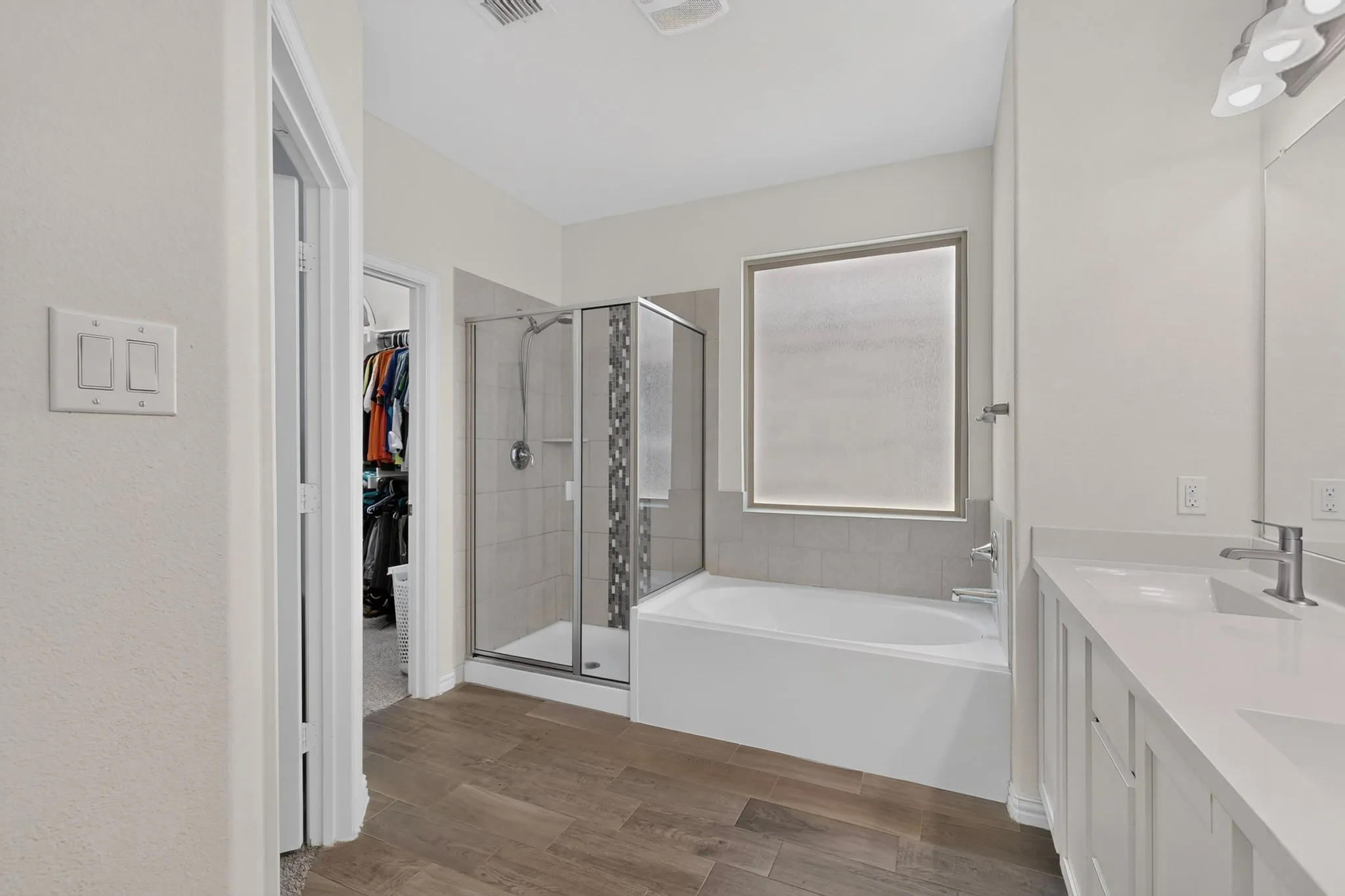 Bathroom featuring wood-finish flooring, a double vanity with white countertops, a built-in tub, and a glass-enclosed shower with tile surround