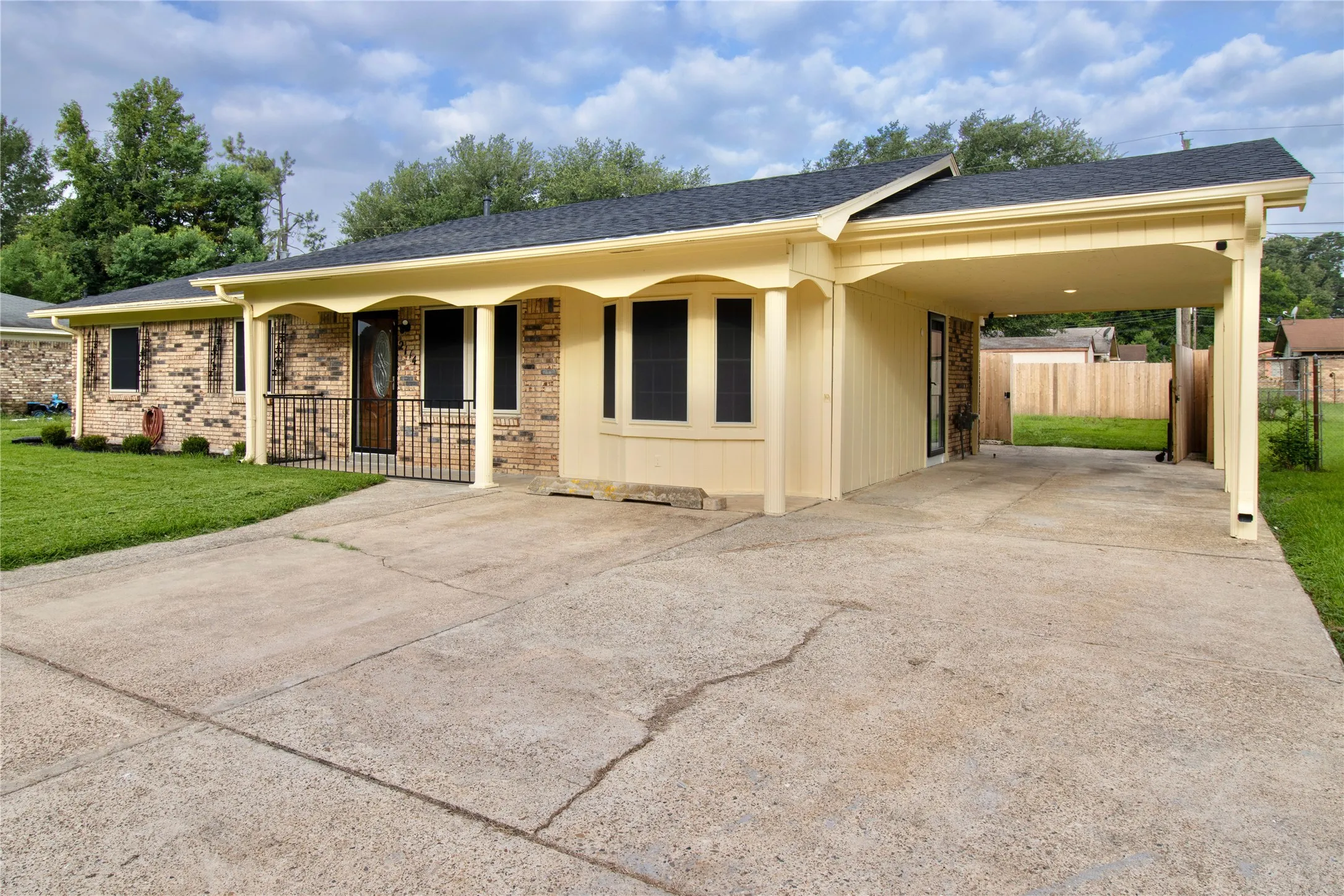 Brick facade residence featuring an attached carport, covered front entry, dark shingle roof, bay window, and manicured front lawn