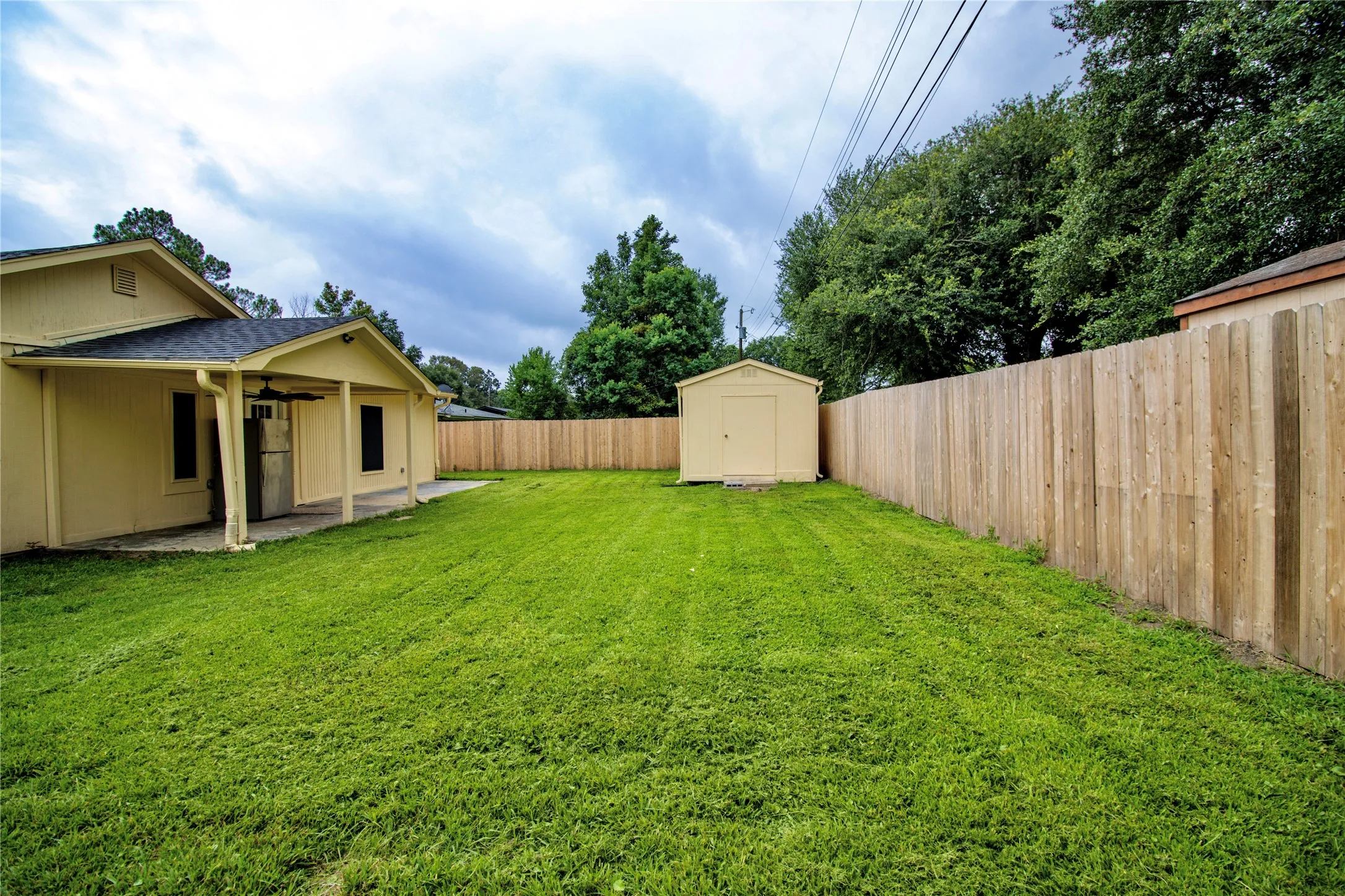 Expansive backyard featuring a lush green lawn, full wood privacy fencing, a covered patio with a ceiling fan, and a detached storage shed