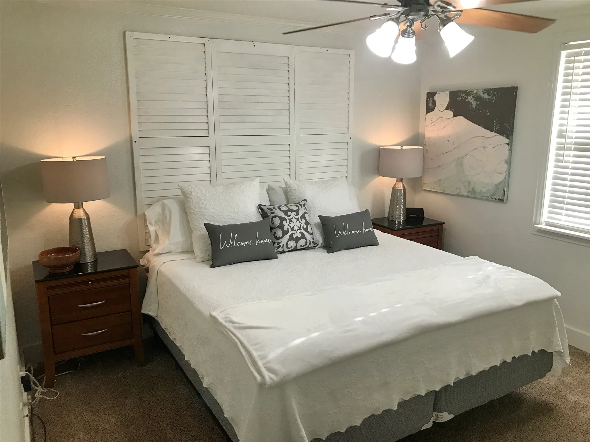 Bedroom featuring a white louvered headboard, textured neutral carpeting, a ceiling fan with integrated lighting, and a window with horizontal blinds