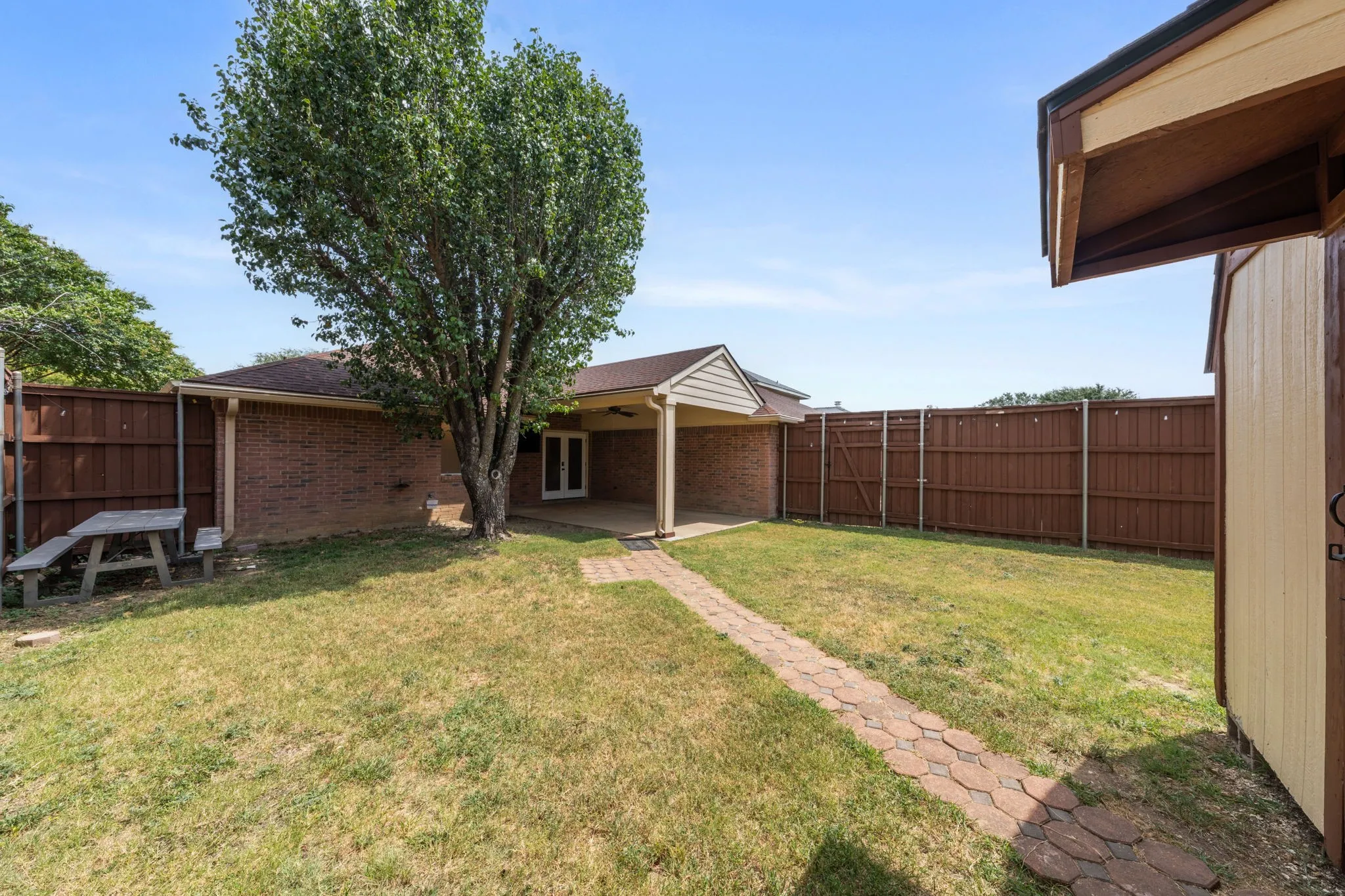Expansive backyard featuring a red brick property exterior, a covered patio, a privacy fence, a paved walkway, and a detached storage shed