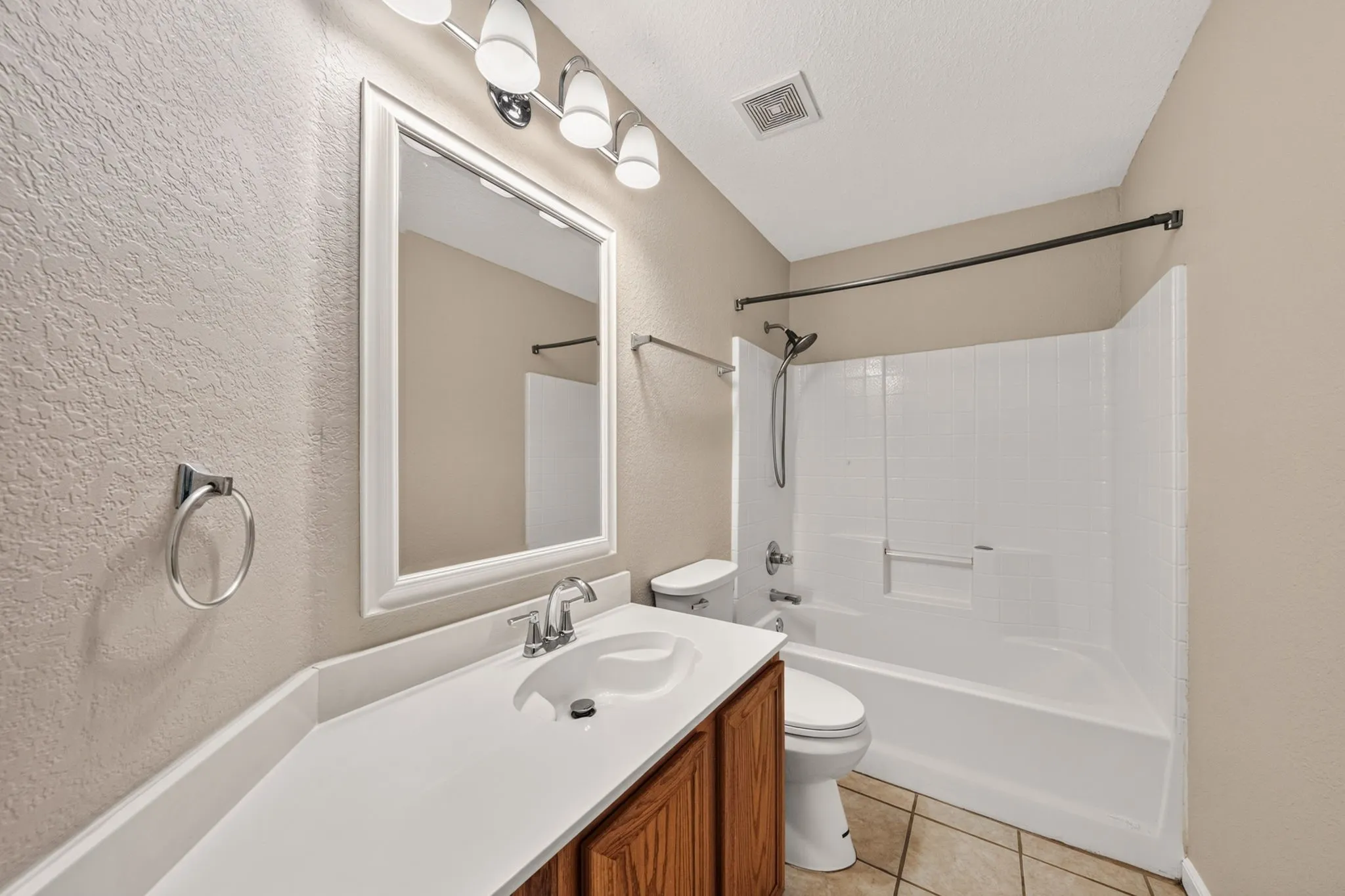 Bright and functional bathroom featuring a wood-finish vanity with dual-sink countertop, framed mirror with integrated lighting, ceramic tile flooring, and a white fiberglass tub-shower combination with glass enclosure and chrome fixtures