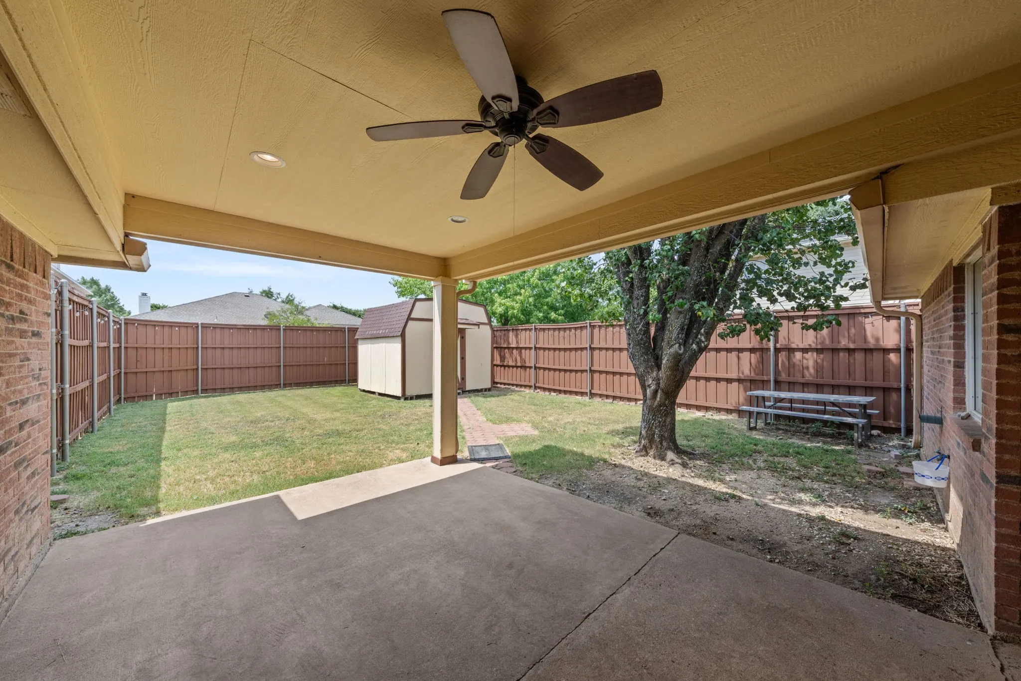 Covered patio with concrete flooring and ceiling fan opens to a spacious fenced backyard featuring mature tree, manicured lawn, and brick privacy fencing