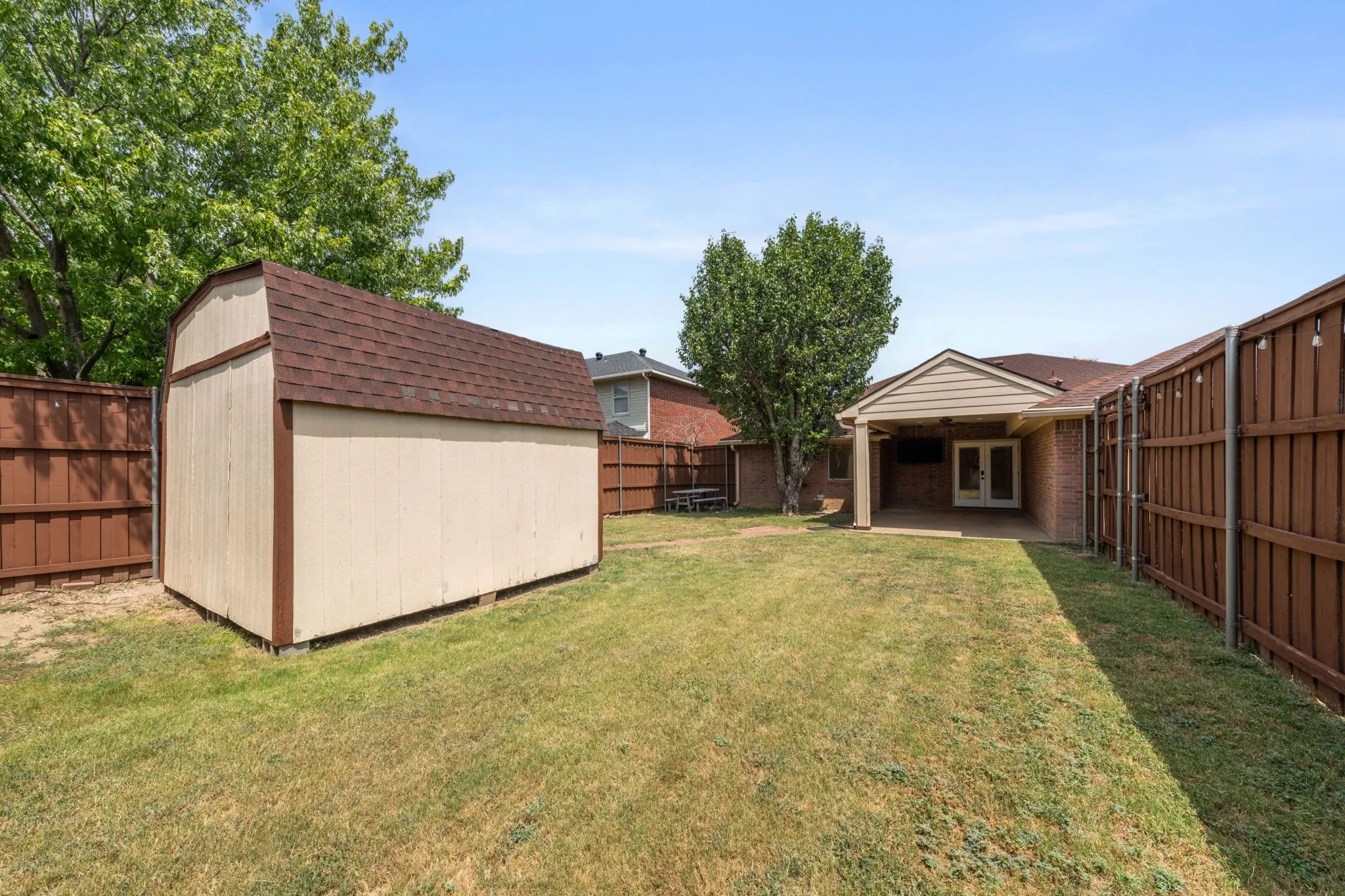 Covered patio with ceiling fan and picnic table seating, overlooking a fenced backyard with mature tree, manicured lawn, and brick home exterior and 12x12 shed!