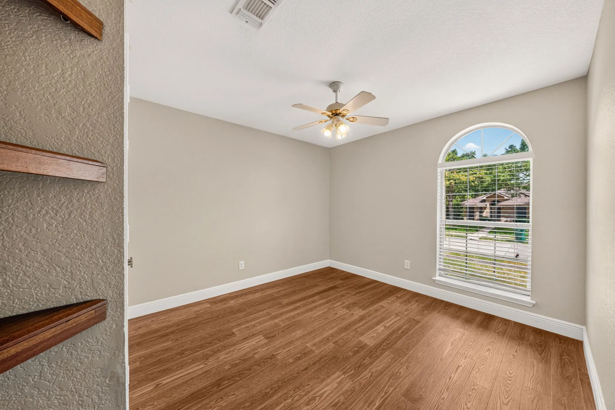 Inviting room with wood-finish flooring, custom shelving an arched window, and a ceiling fan with integrated lighting and cieling fans