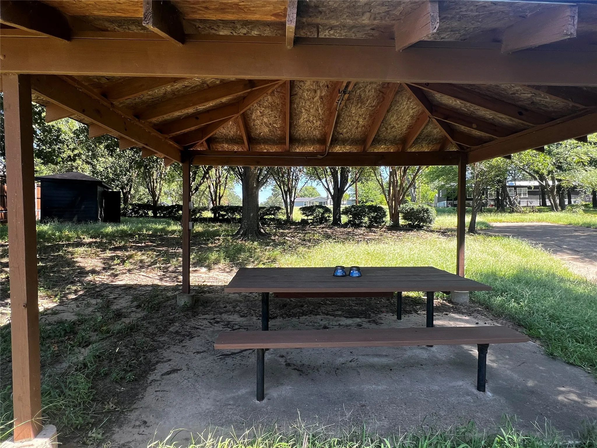 Open-air covered patio with a wood-finish roof and support beams, featuring an integrated picnic table on a concrete pad facing the inland portion of the lots.  The lake is behind the photographer.   The rest of the land goes beyond the hedges up to the blue house in the background.  Boat ramp is to the right.