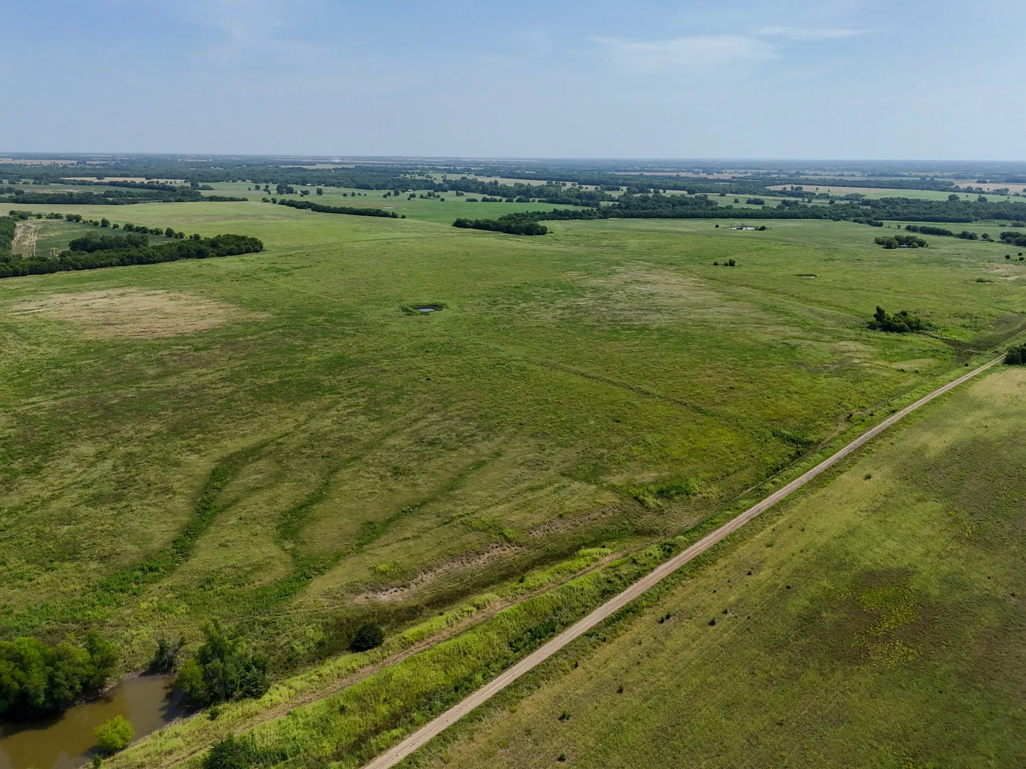 Expansive green acreage with diverse topography, featuring a dirt road, scattered trees, and a visible pond