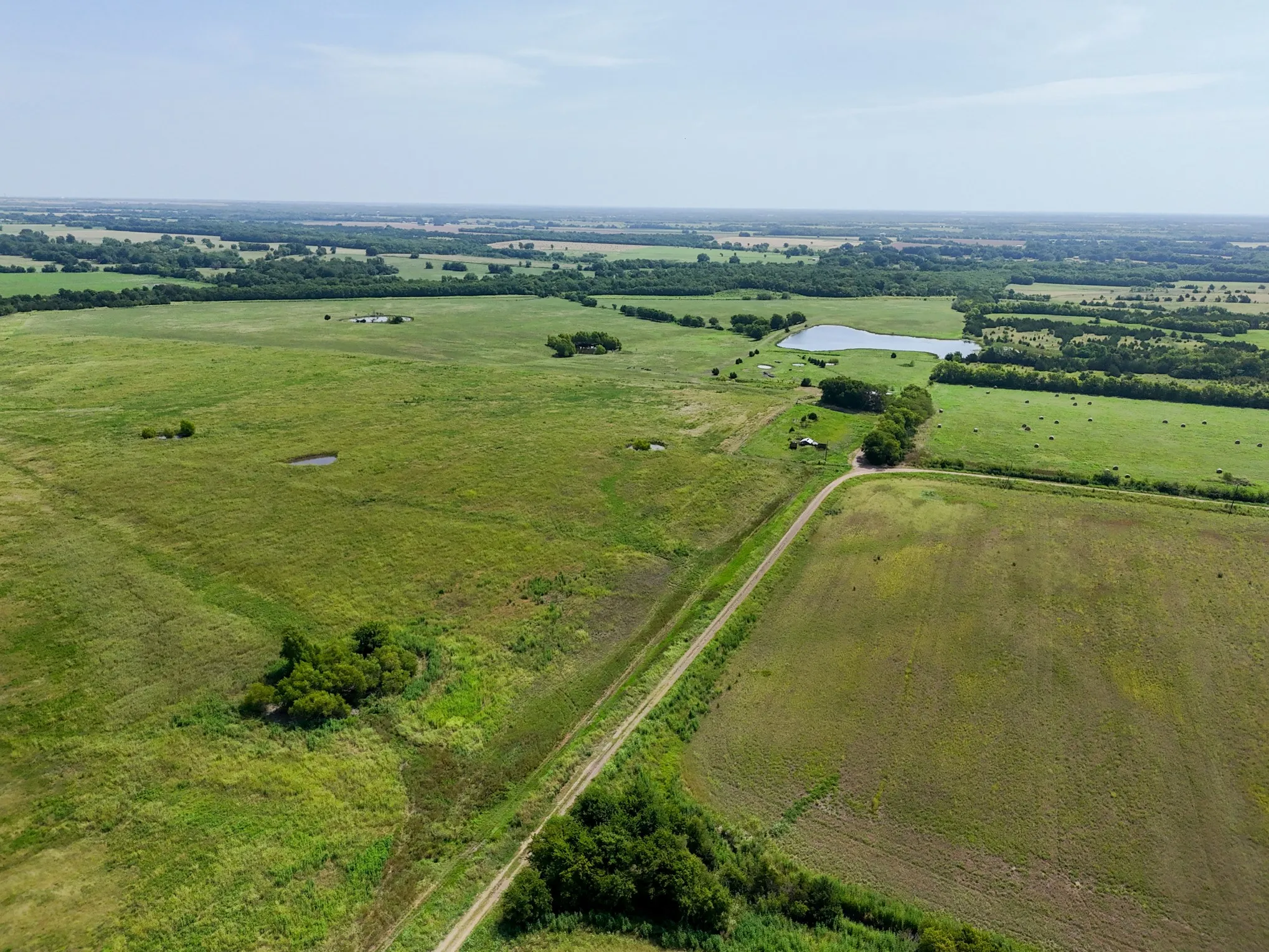 Expansive rural landscape featuring a large body of water, green pastures, scattered trees, and a visible track road