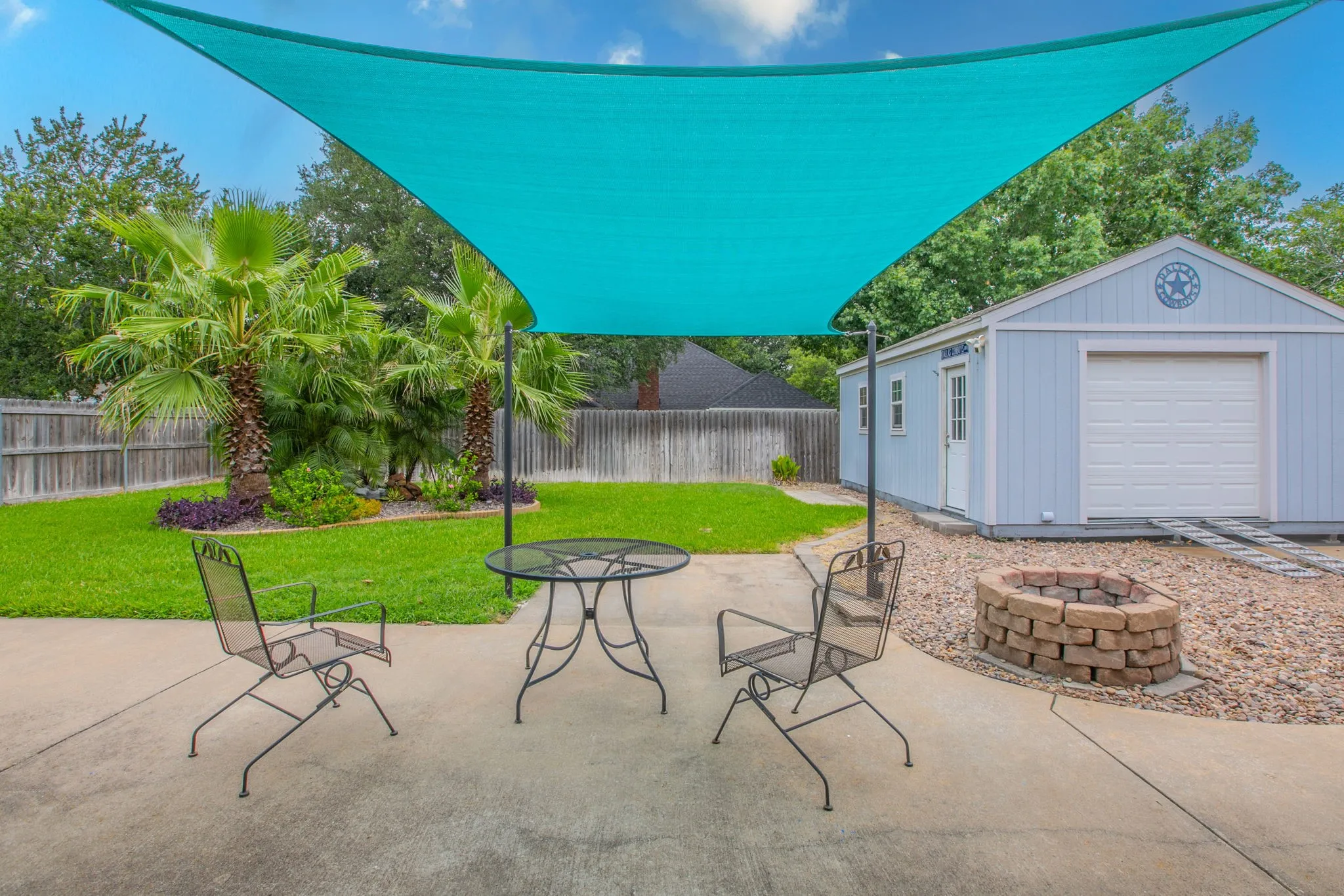 Expansive backyard featuring a concrete patio with a shade sail, a stone fire pit, and a separate light blue utility building with a roll-up garage door and an entry door