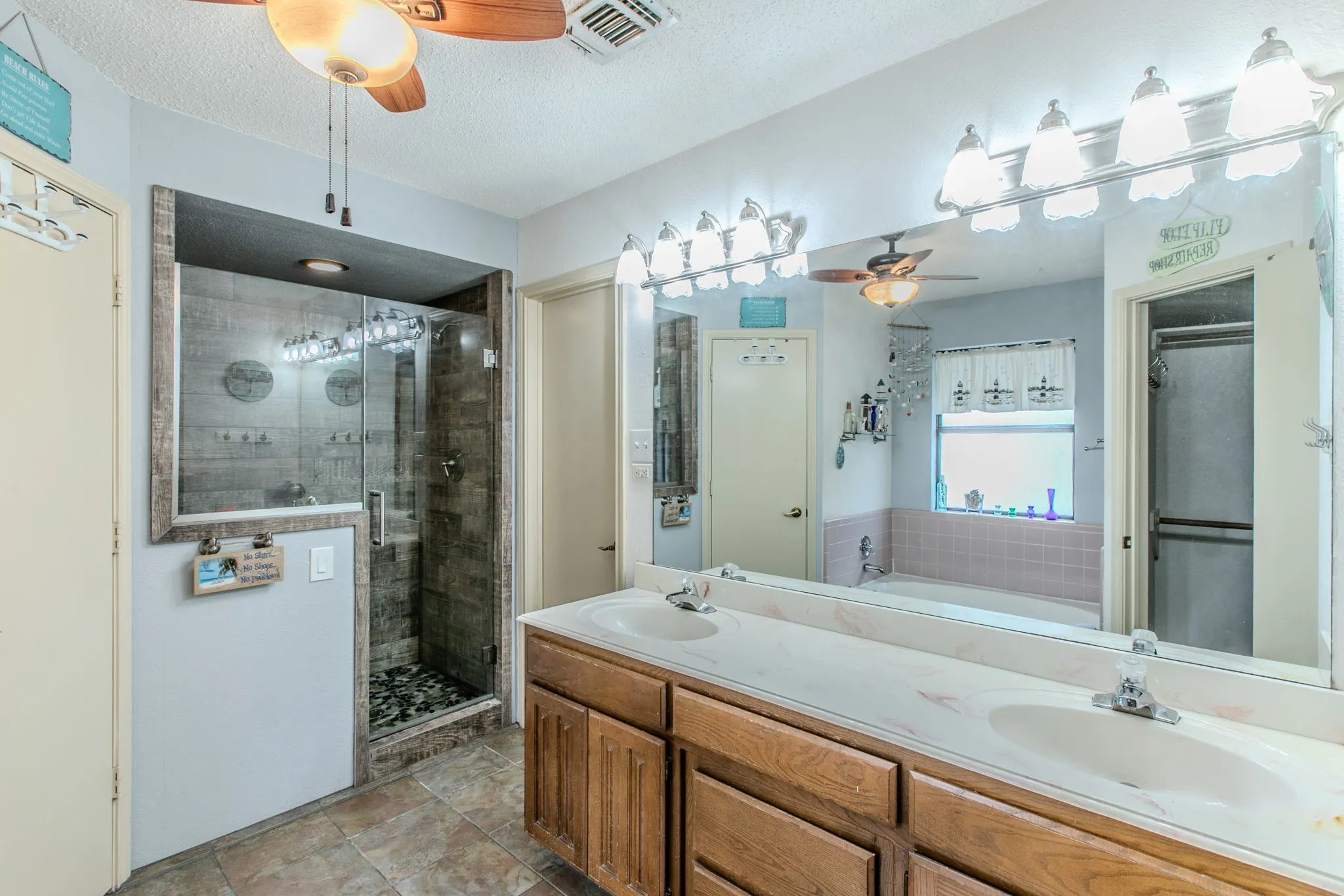 Bathroom featuring a dual vanity with laminate countertops, a glass-enclosed shower with tile surround and pebble floor, and a built-in bathtub with tiled wainscoting