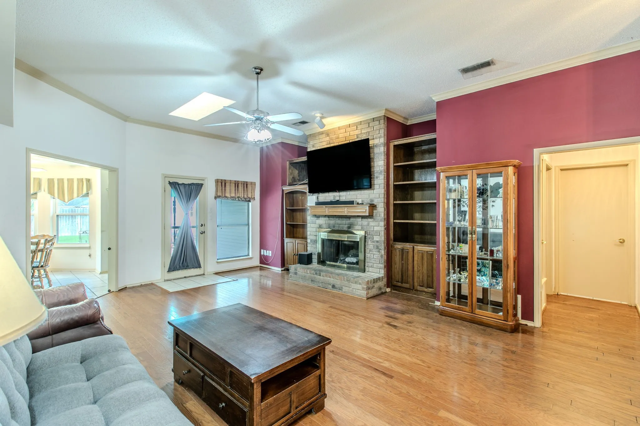 Living area featuring wood-finish flooring, a brick fireplace with mantel, built-in shelving, vaulted ceilings, and a skylight
