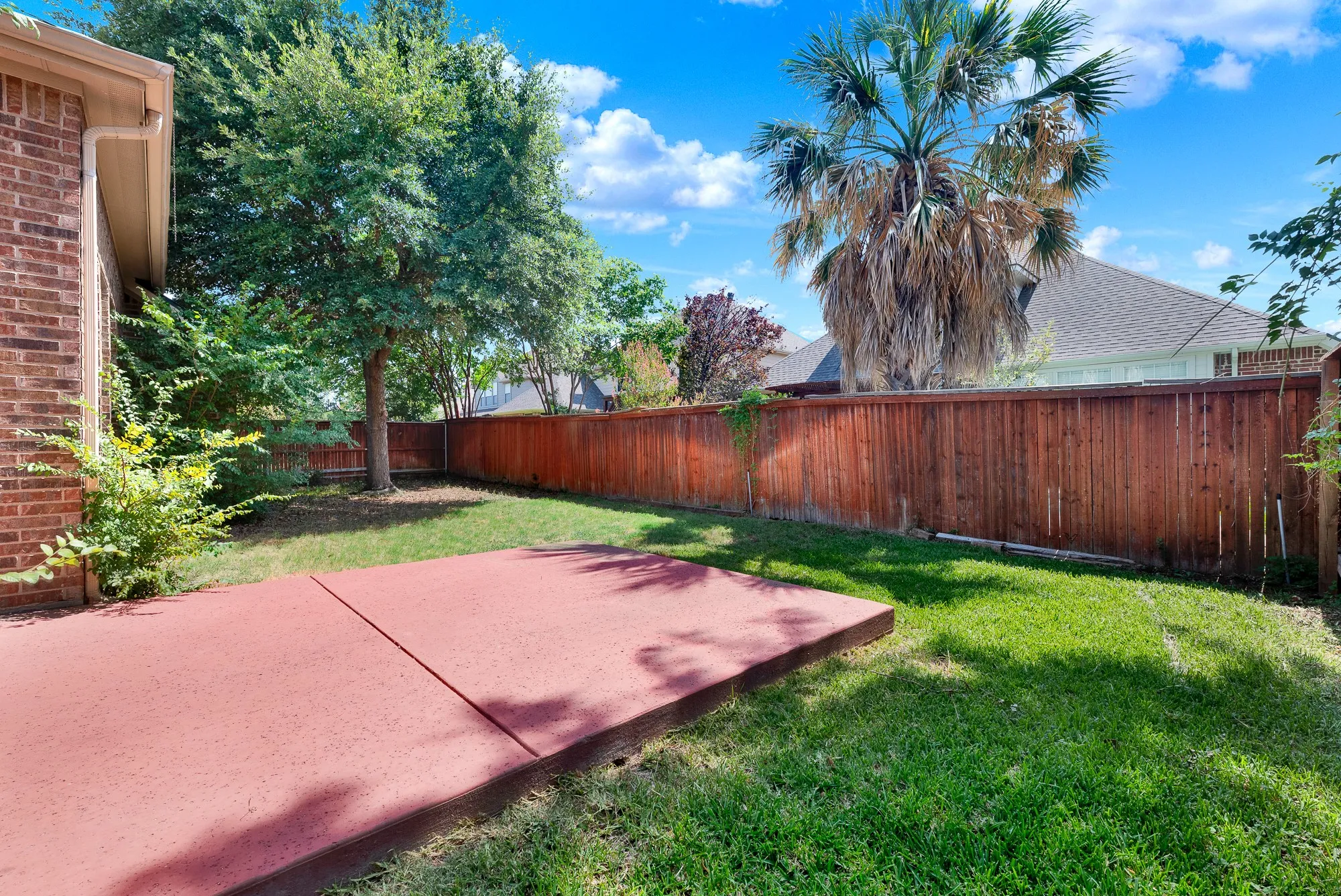 Landscaped backyard featuring a red concrete patio, wood privacy fencing, and mature trees