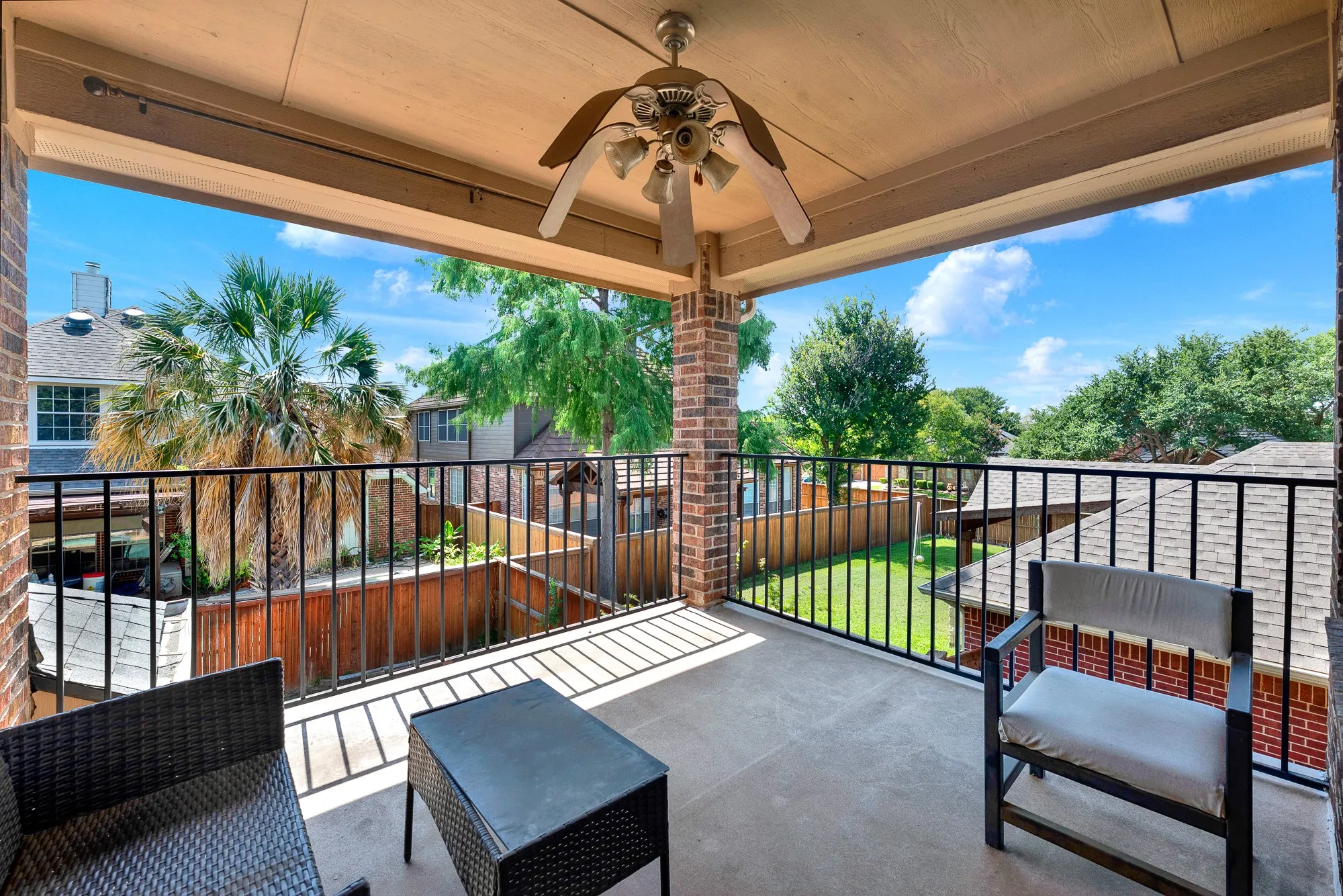 Covered balcony with a ceiling fan, brick support columns, and black metal railings overlooking a backyard with green grass and mature trees