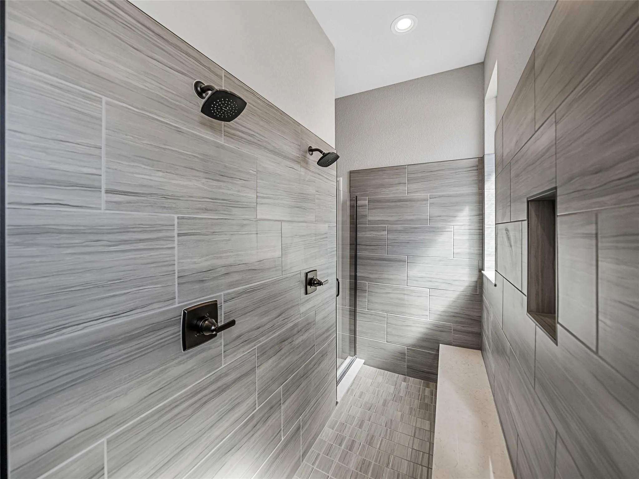Spacious shower enclosure featuring large format stone-look wall tiles, dual matte black shower heads, a built-in shower niche, a recessed ceiling light, and a stone bench