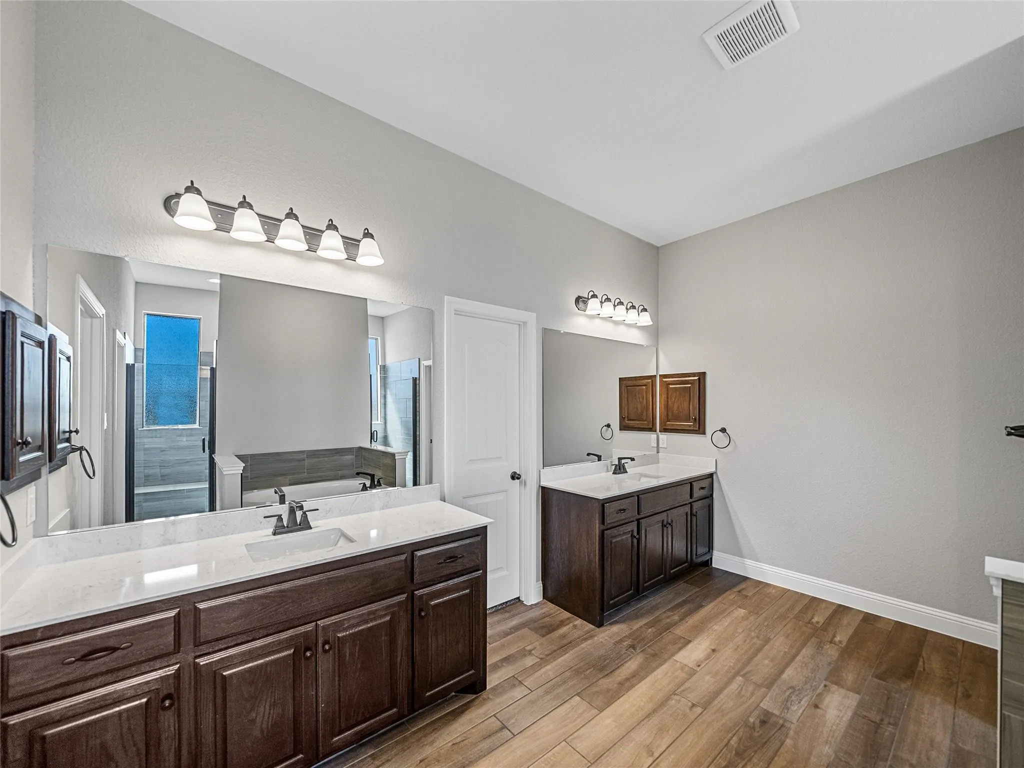 Spacious bathroom featuring dual vanities with white countertops, dark wood cabinetry, wood-finish flooring, and individual vanity lighting fixtures