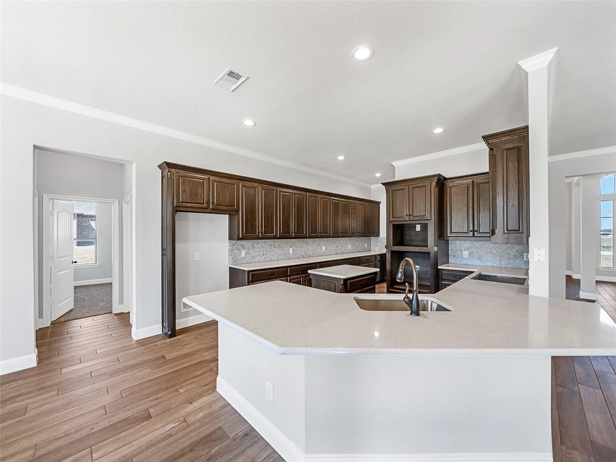 Open-concept kitchen featuring an expansive island with an undermount sink and gooseneck faucet, abundant dark wood cabinetry, light-toned countertops, and wood-finish flooring