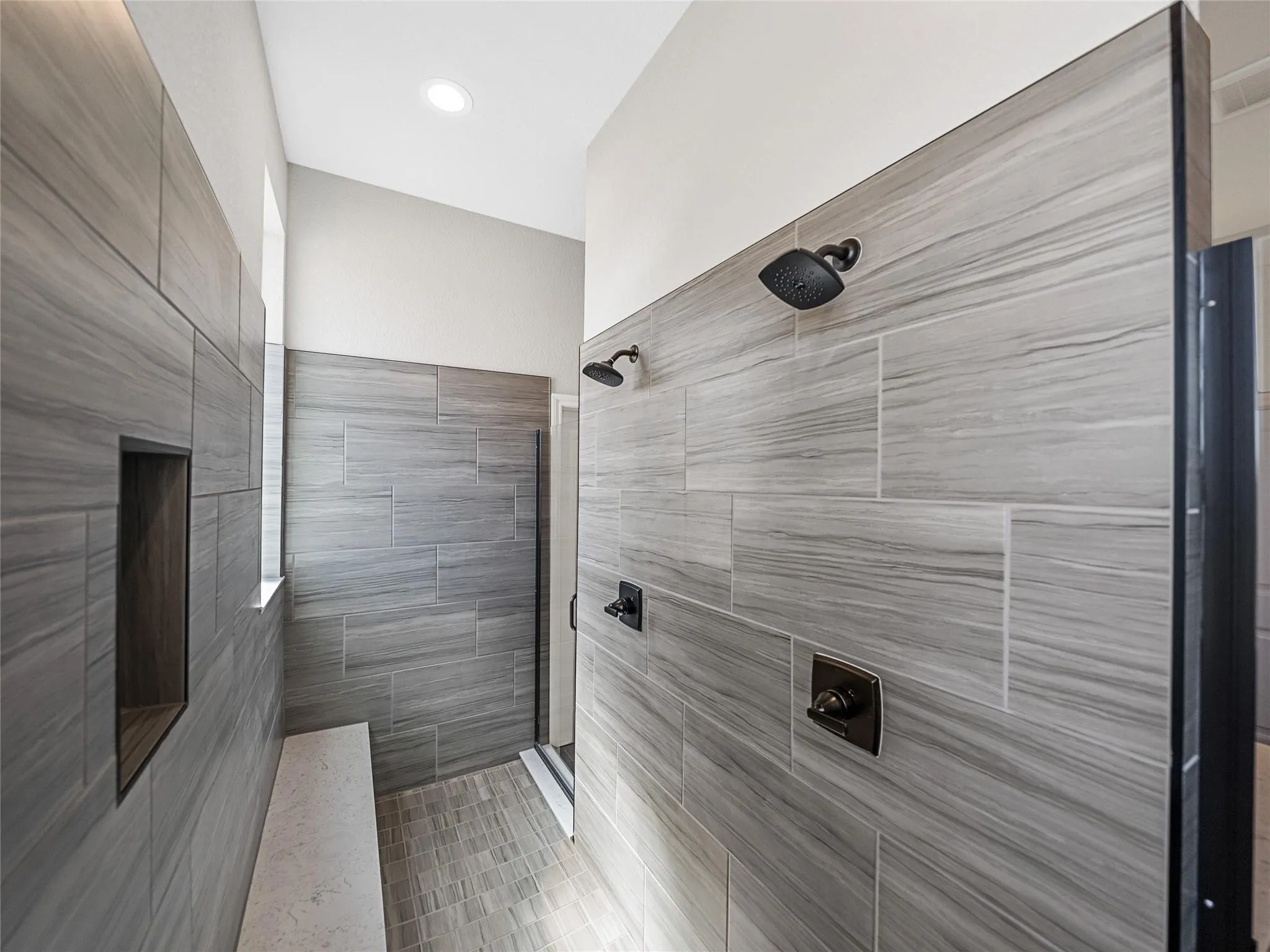 Spacious shower enclosure featuring large format stone-look tile, dual shower heads with matte black fixtures, recessed lighting, a built-in shower bench, and a tiled niche