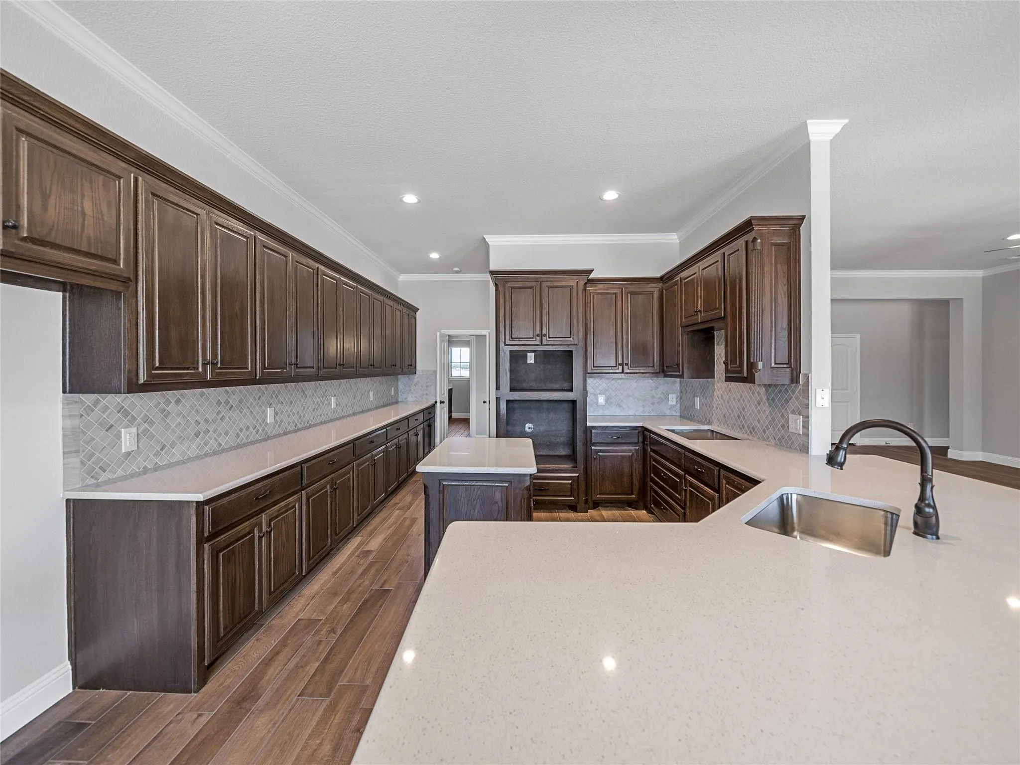 Spacious kitchen featuring extensive dark wood cabinetry, light-toned countertops, a mosaic tile backsplash, wood-finish flooring, and a large island with an undermount sink and gooseneck faucet