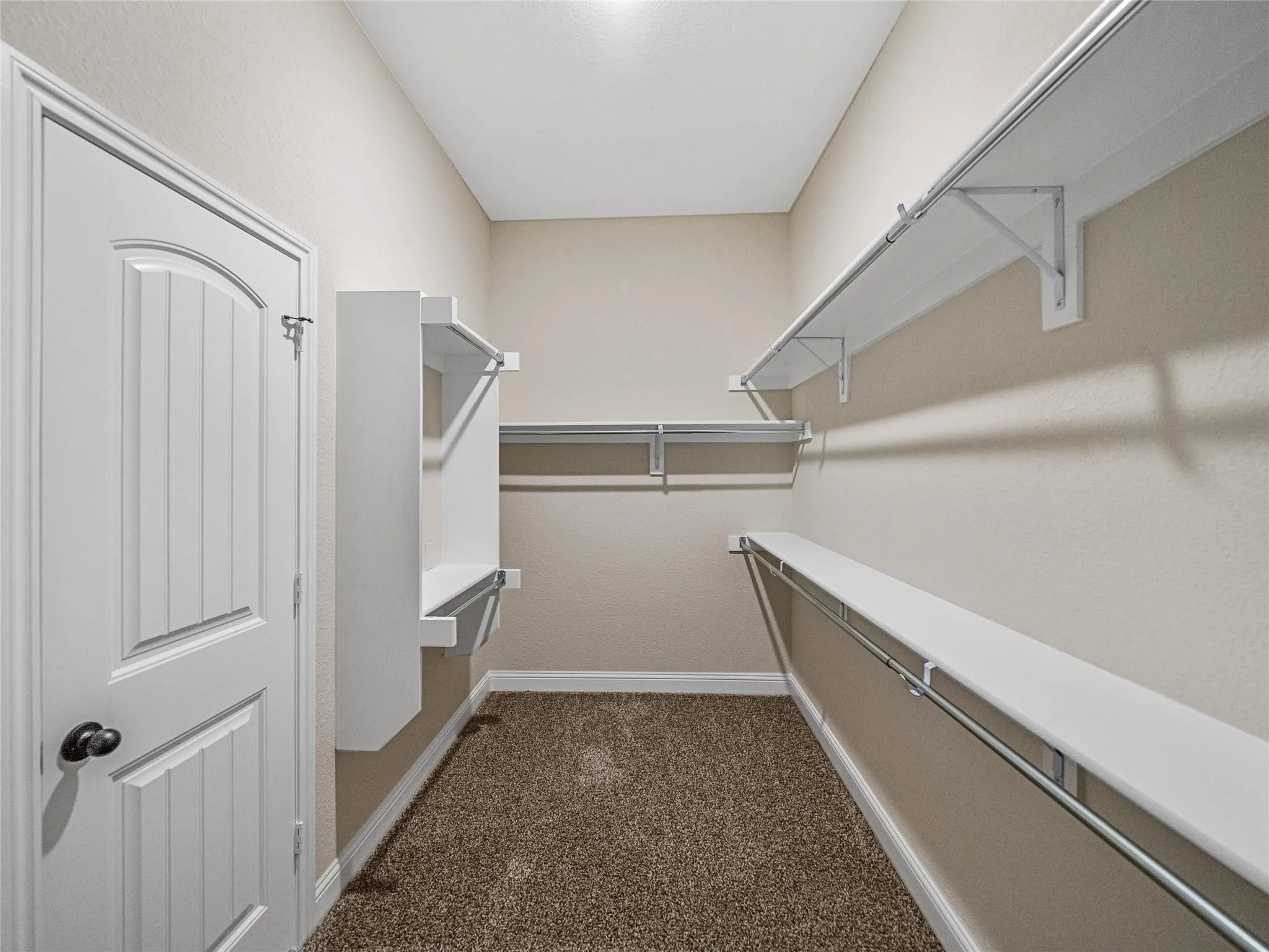 Walk-in closet featuring built-in shelving, hanging rods, an arched panel door, light-colored walls, and brown carpet flooring