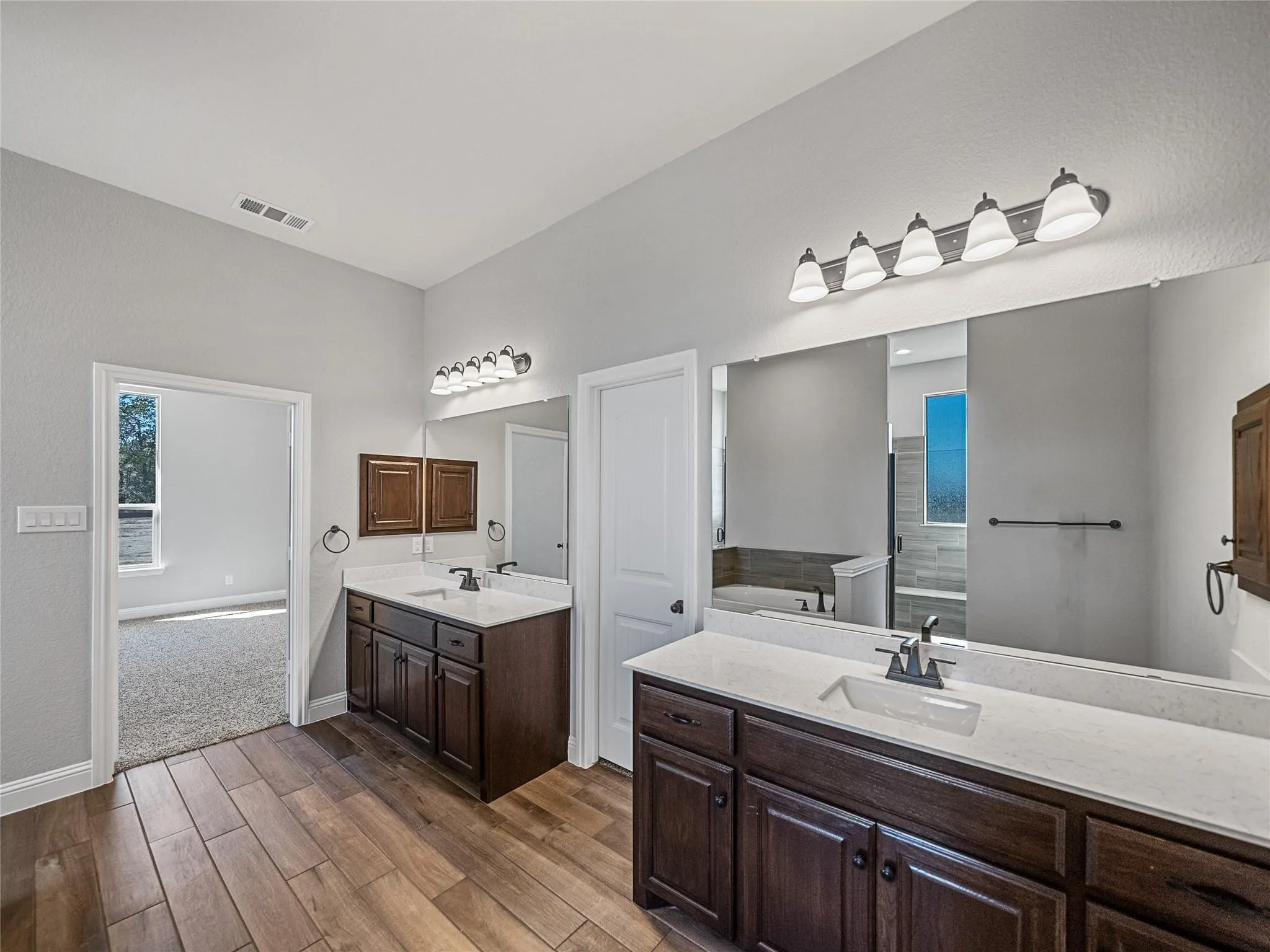 Spacious bathroom featuring wood-finish flooring, dual dark wood vanities with light countertops, and individual vanity lighting