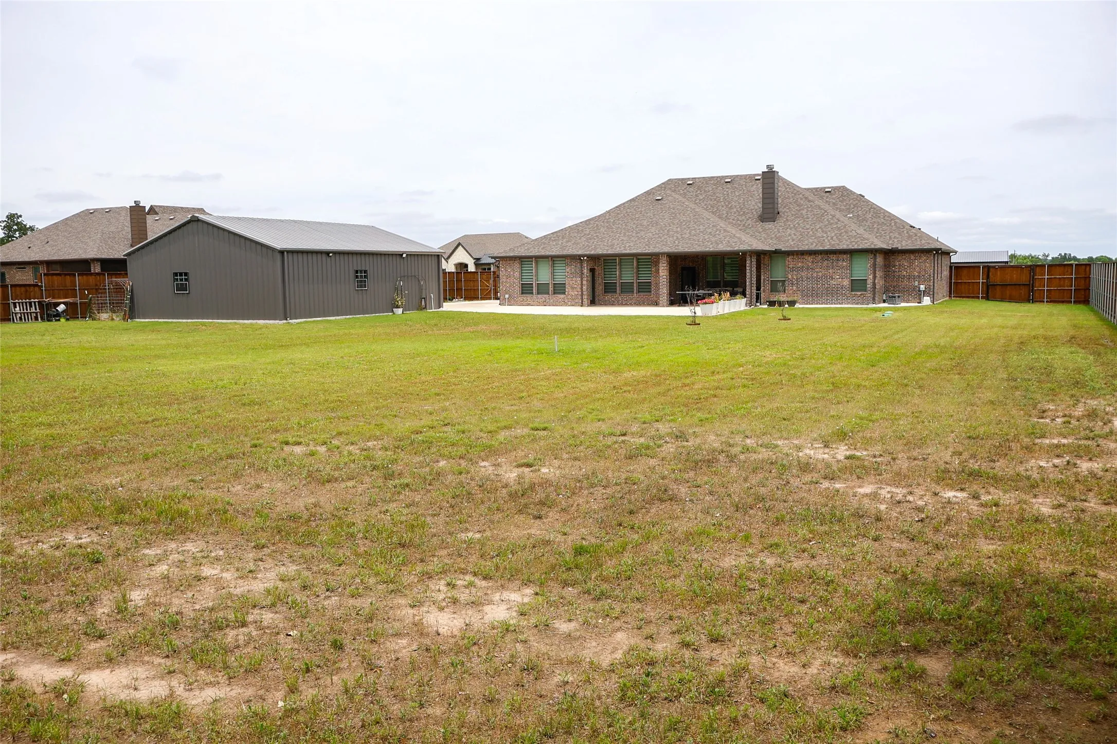 Expansive rear yard featuring a brick-clad residence with a covered patio, a separate metal utility building, and a wood privacy fence