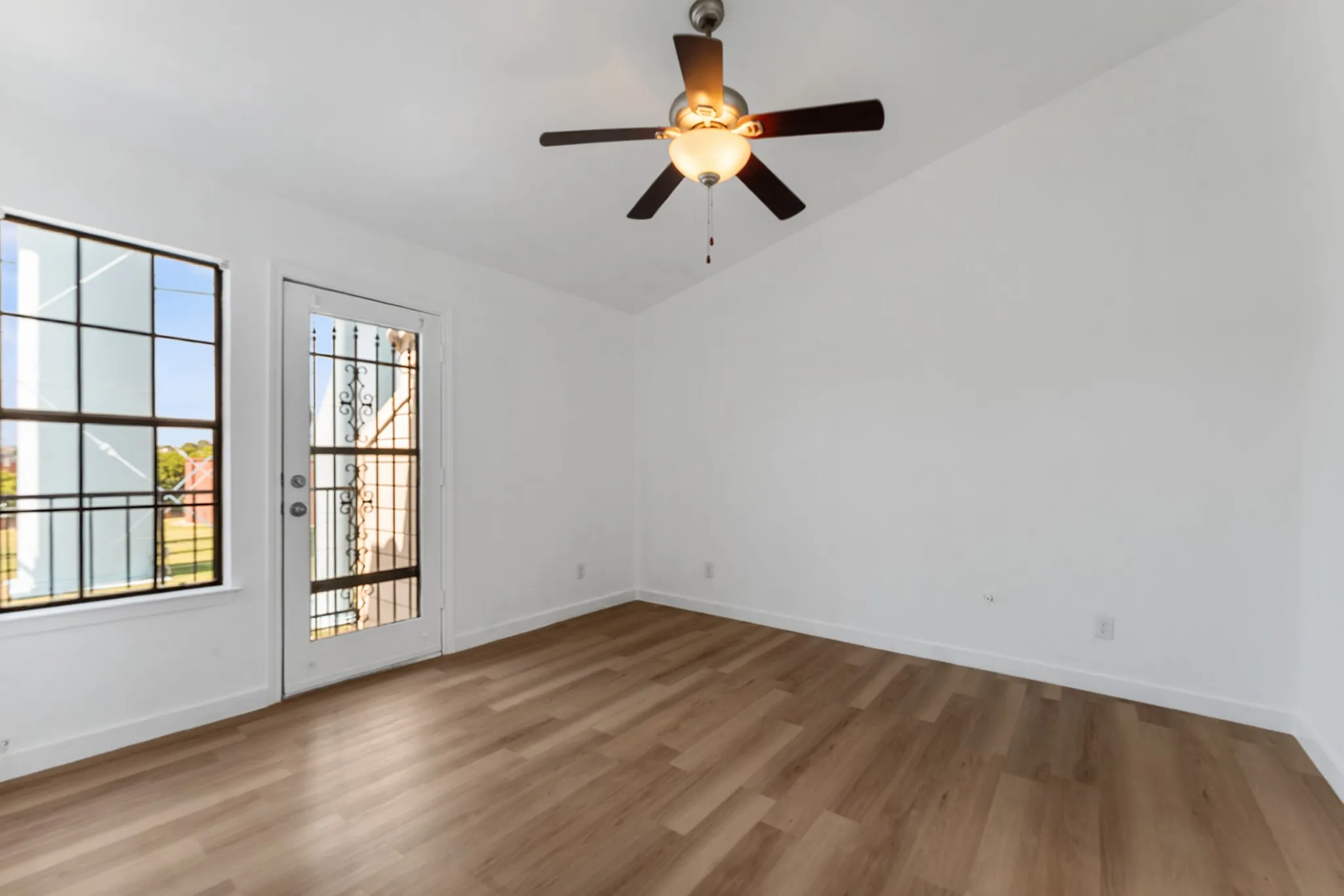Spacious room featuring wood-finish flooring, a contemporary ceiling fan with integrated lighting, a large multi-pane window, a glass-panel door with decorative security grate, and crisp white walls