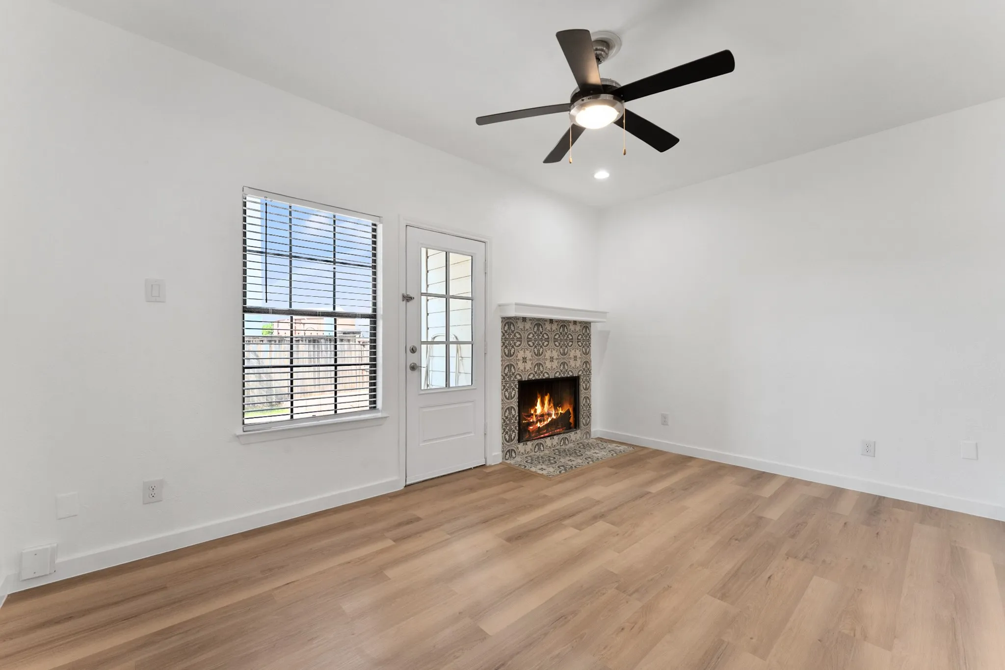 Living area featuring wood-finish flooring, a decorative tile fireplace surround, a multi-pane window with blinds, a white paneled door with glass inserts, and a ceiling fan with integrated lighting