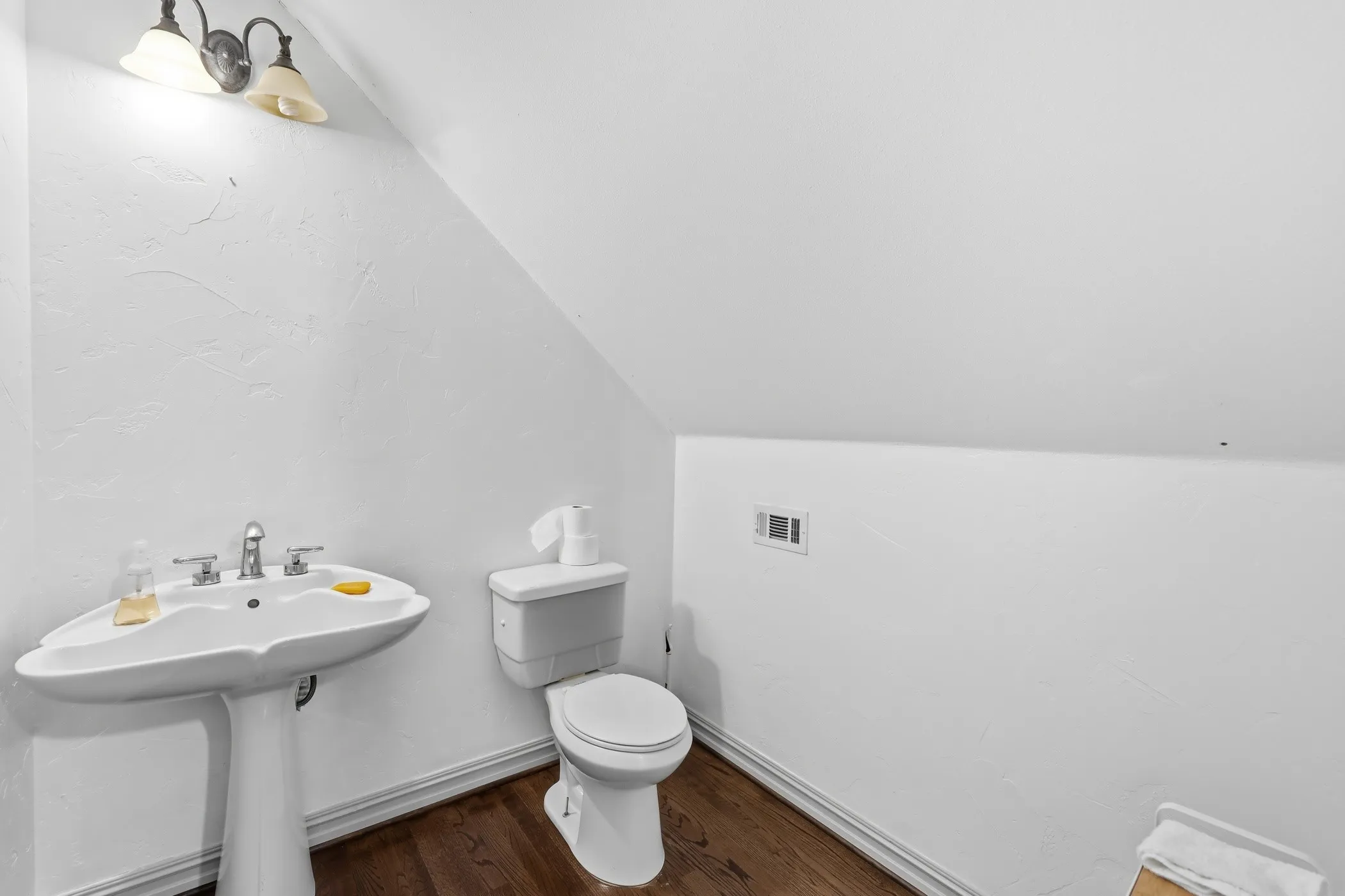 Pedestal sink with chrome faucet, complemented by a white toilet, wood-finish flooring, and a wall-mounted light fixture