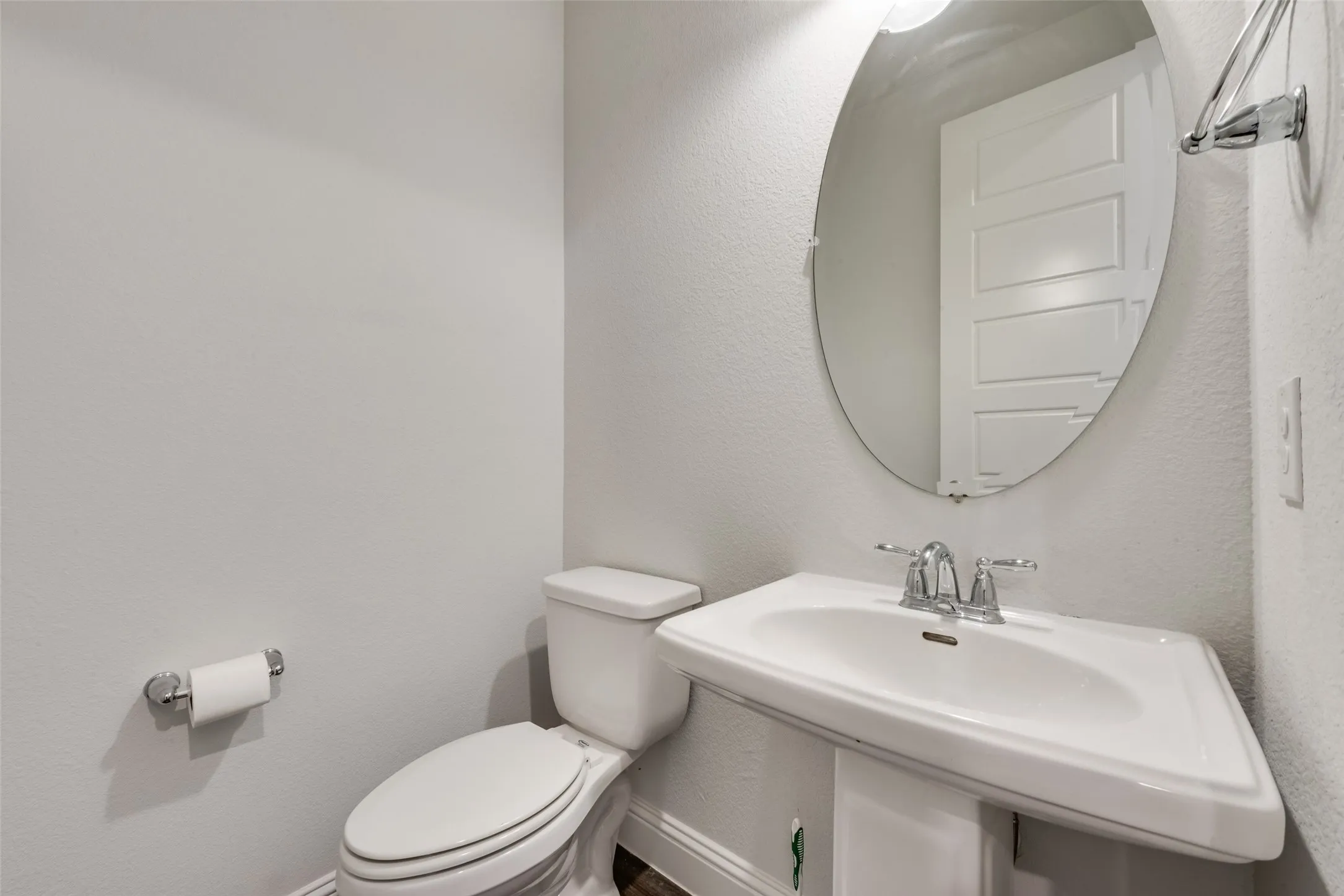 Pedestal sink with chrome fixtures and an oval mirror, complemented by a white toilet, chrome toilet paper holder, and dark wood-finish flooring