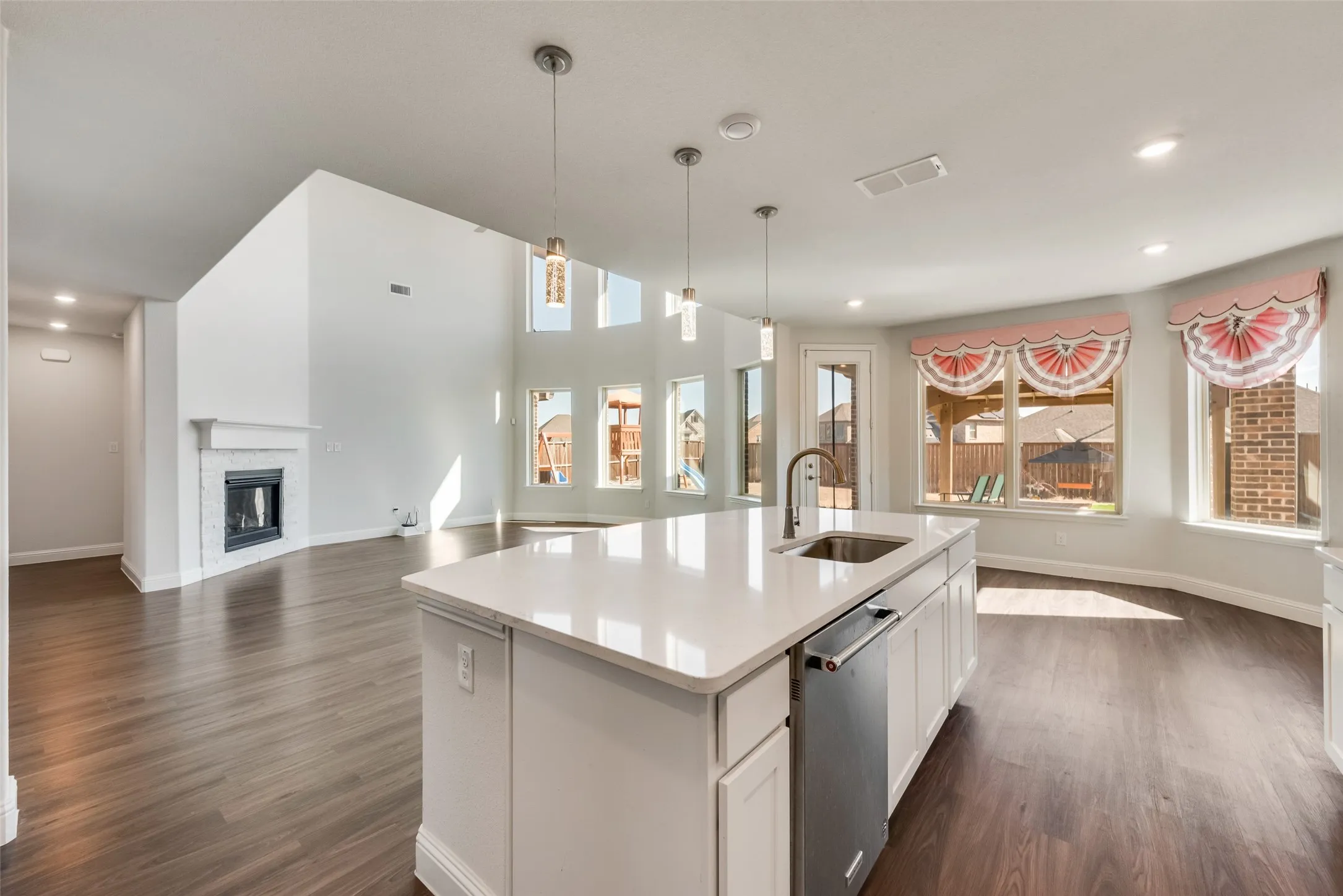 Open-concept living area featuring a kitchen island with sink and integrated dishwasher, wood-finish flooring, a fireplace with a white mantel, and vaulted ceilings with multiple windows