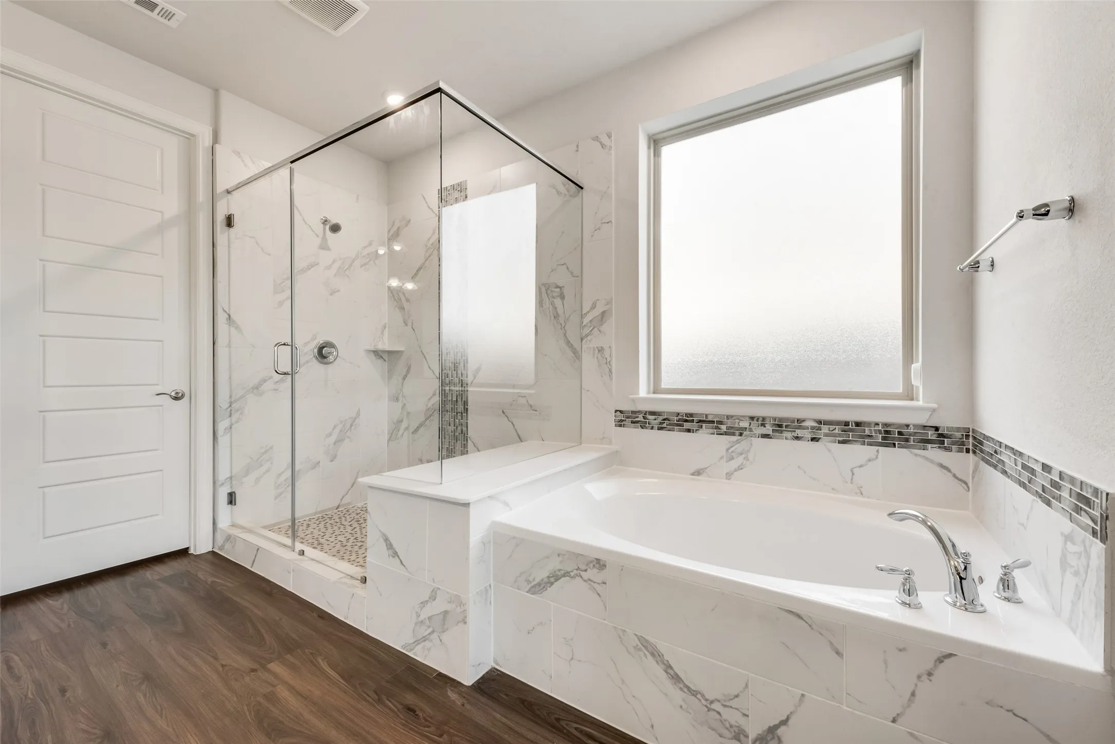 Bathroom featuring a frameless glass shower enclosure with pebble floor, a built-in tub with chrome fixtures, and white and gray marbled tile surround