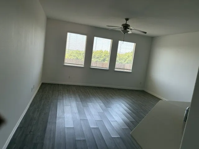 Bright room featuring three windows with blinds, wood-finish flooring, a ceiling fan, and white walls