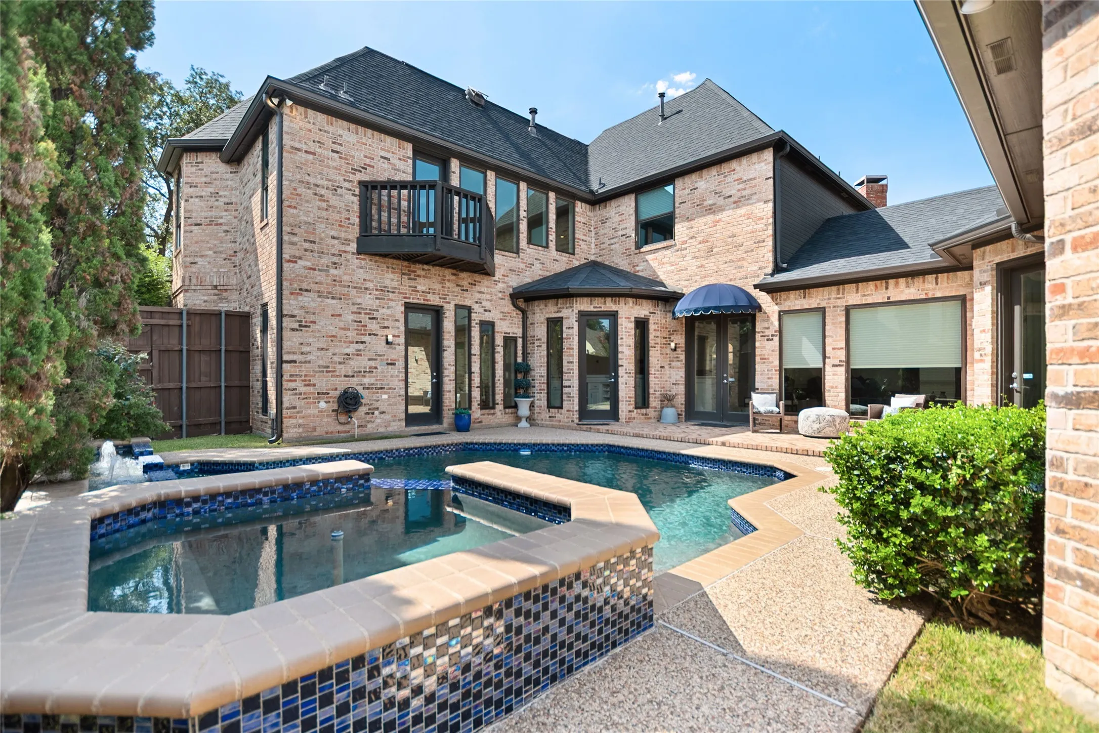 Brick exterior residence featuring a multi-level pool with integrated spa, an elevated balcony, multiple access doors, and an exposed aggregate patio