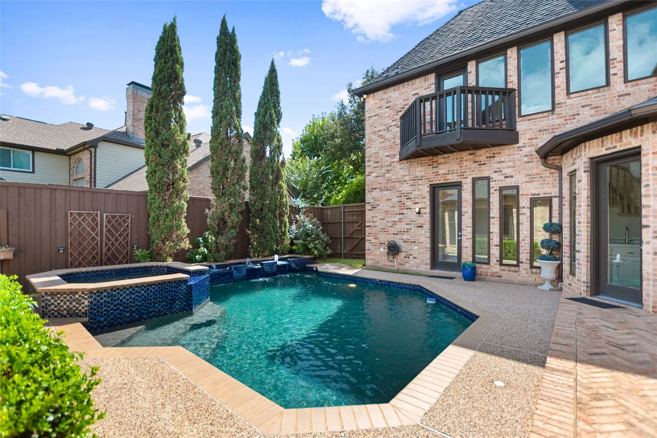 Expansive backyard featuring a geometric pool with waterfalls, an integrated spa with blue tile accents, a private patio area, and mature evergreen trees