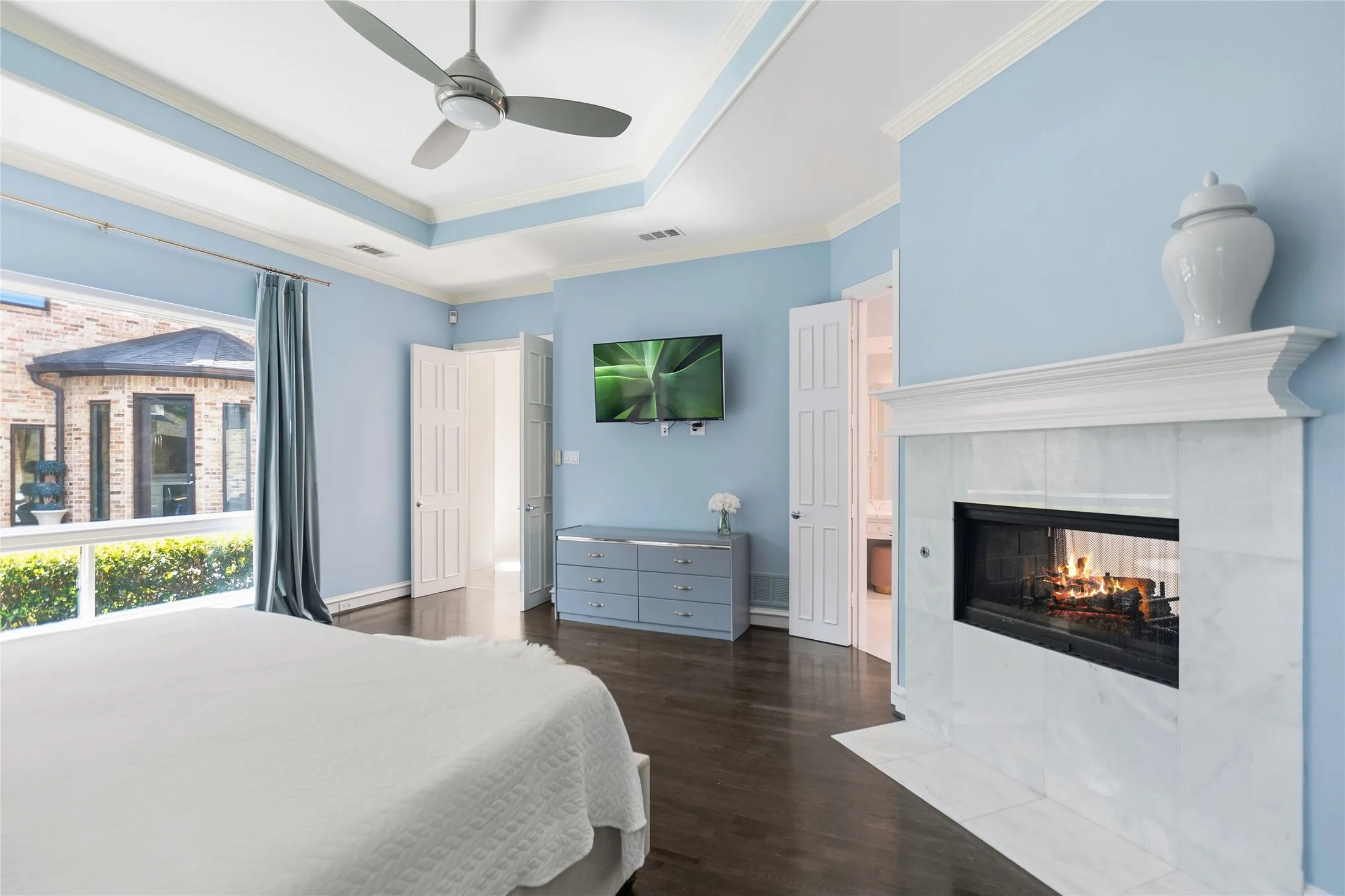 Bedroom featuring a fireplace with a white mantel and tile surround, dark wood-finish flooring, a tray ceiling with a ceiling fan, and large windows with exterior foliage