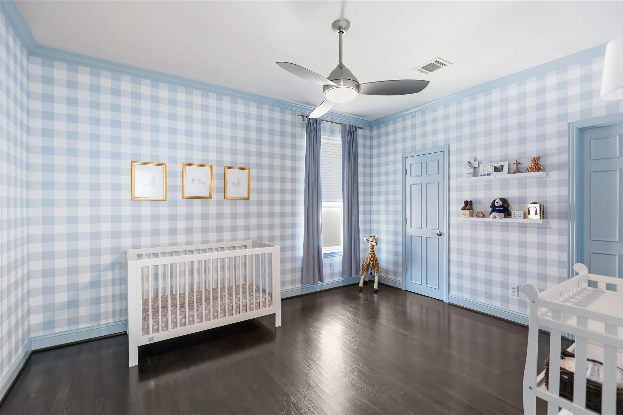 Room with gingham pattern wallpaper, dark wood-finish flooring, two panel doors, an overhead fan with integrated lighting, and window with blinds and curtains