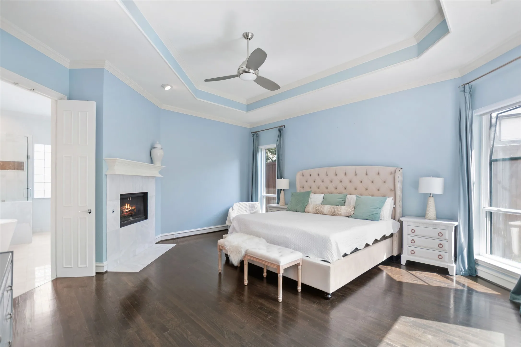 Spacious room featuring dark wood-finish flooring, a white marble fireplace, and a recessed tray ceiling with crown molding